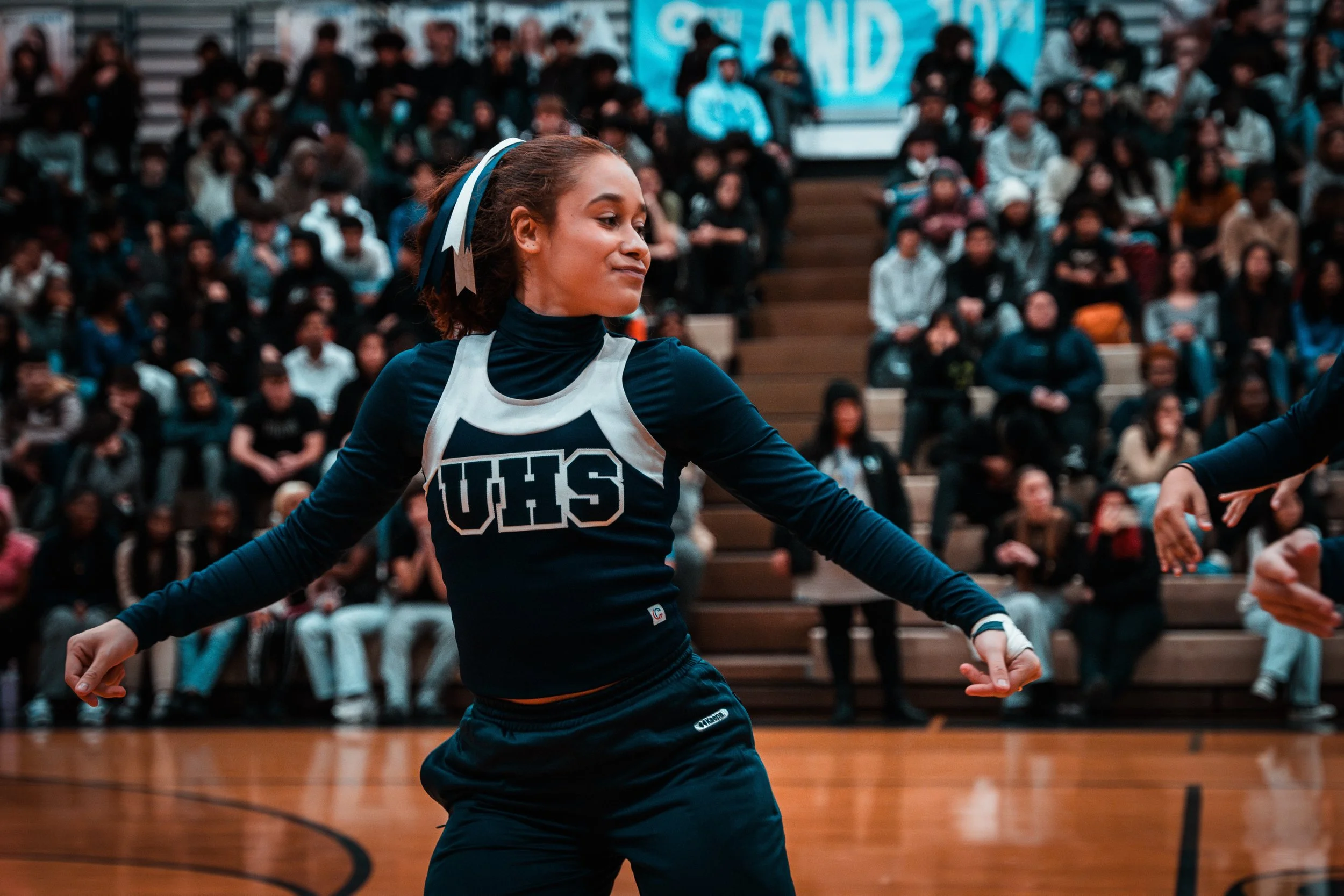 A young woman in a sports uniform with 'UHS' on the front, dancing or performing on a gymnasium floor during a school event, with an audience of students seated in bleachers in the background.