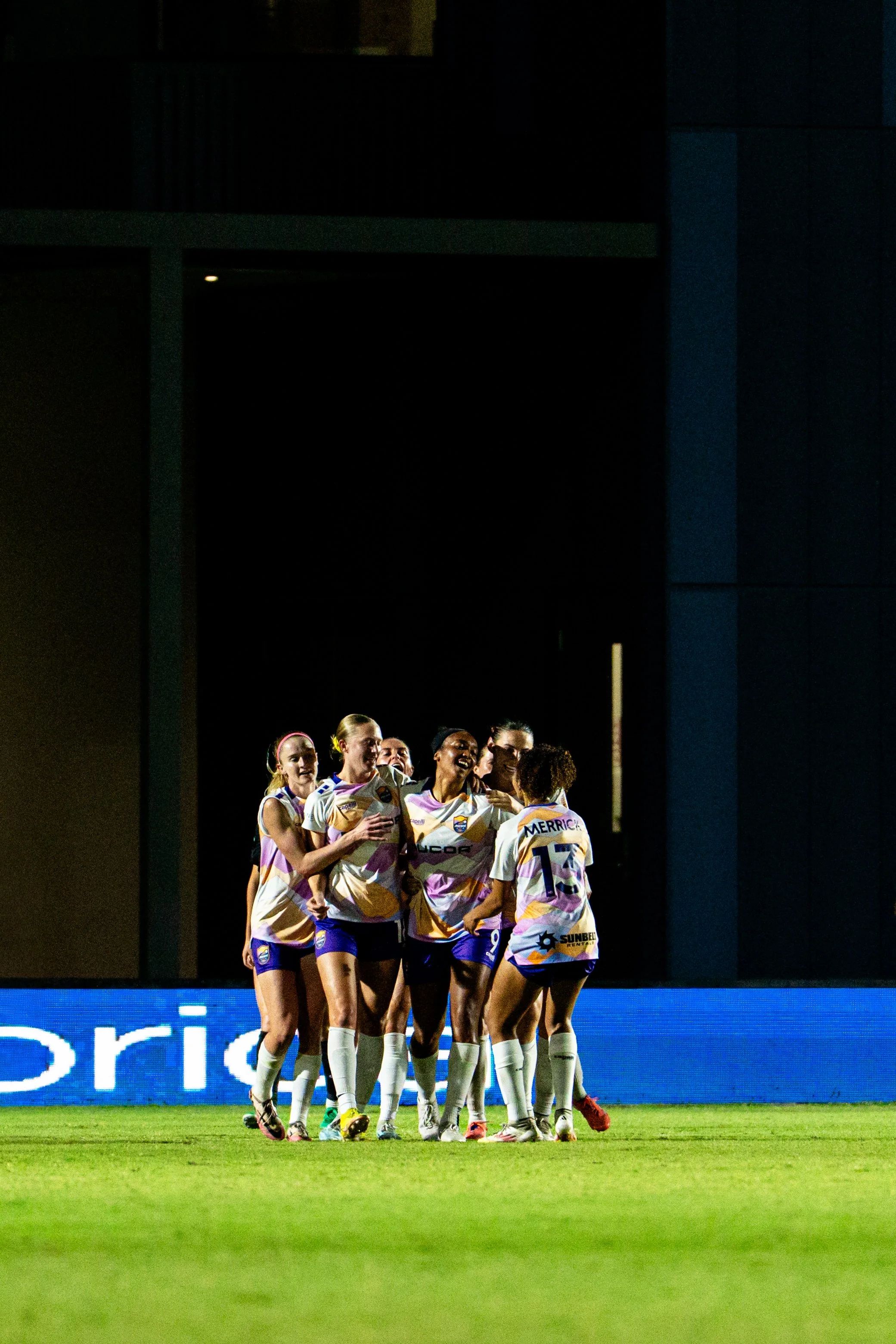 A group of female soccer players celebrating on the field during a match at night. They are wearing white and purple uniforms.