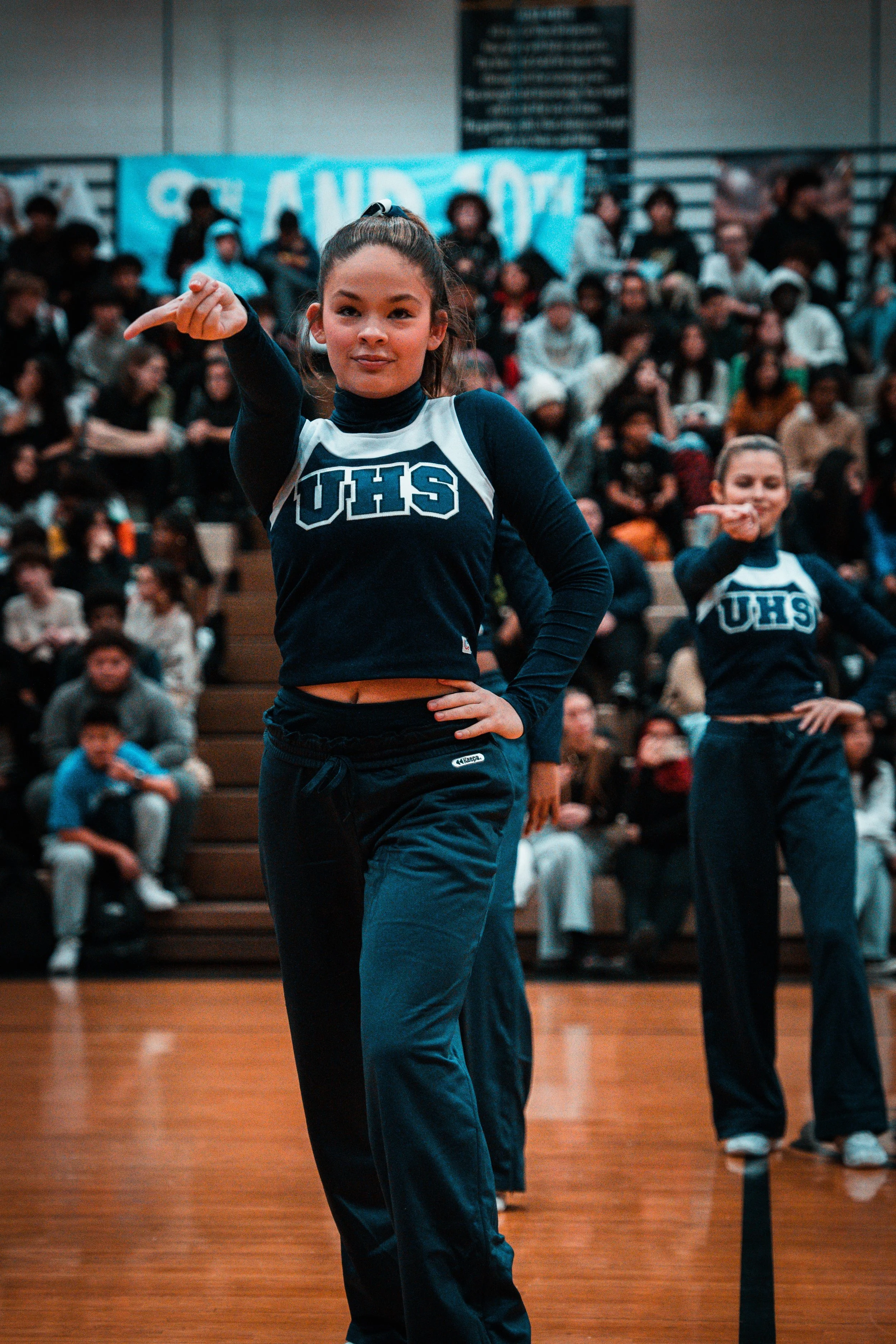 Cheerleader performing a dance routine on a gymnasium floor with a cheering crowd in the background.