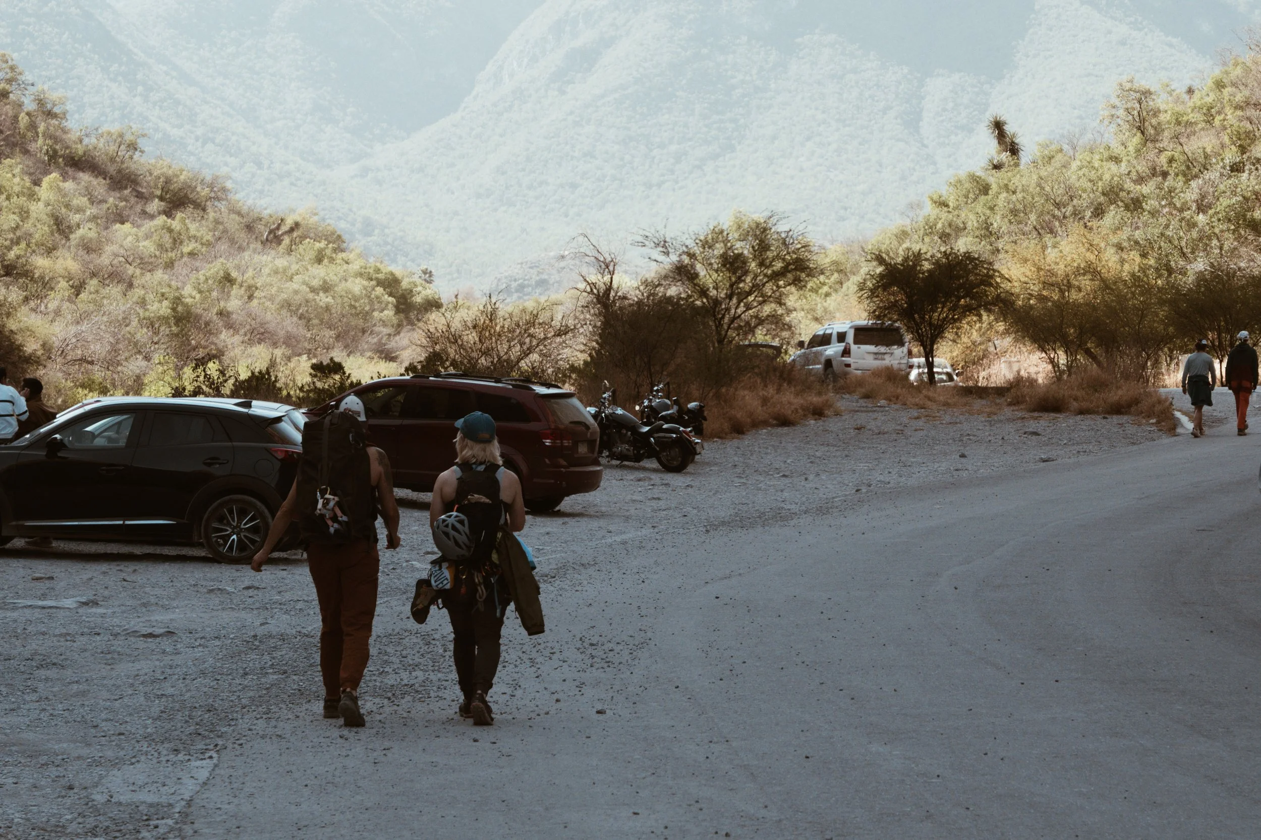 Two hikers walking on a dirt road in a mountainous desert landscape with parked cars and motorcycles on the side.