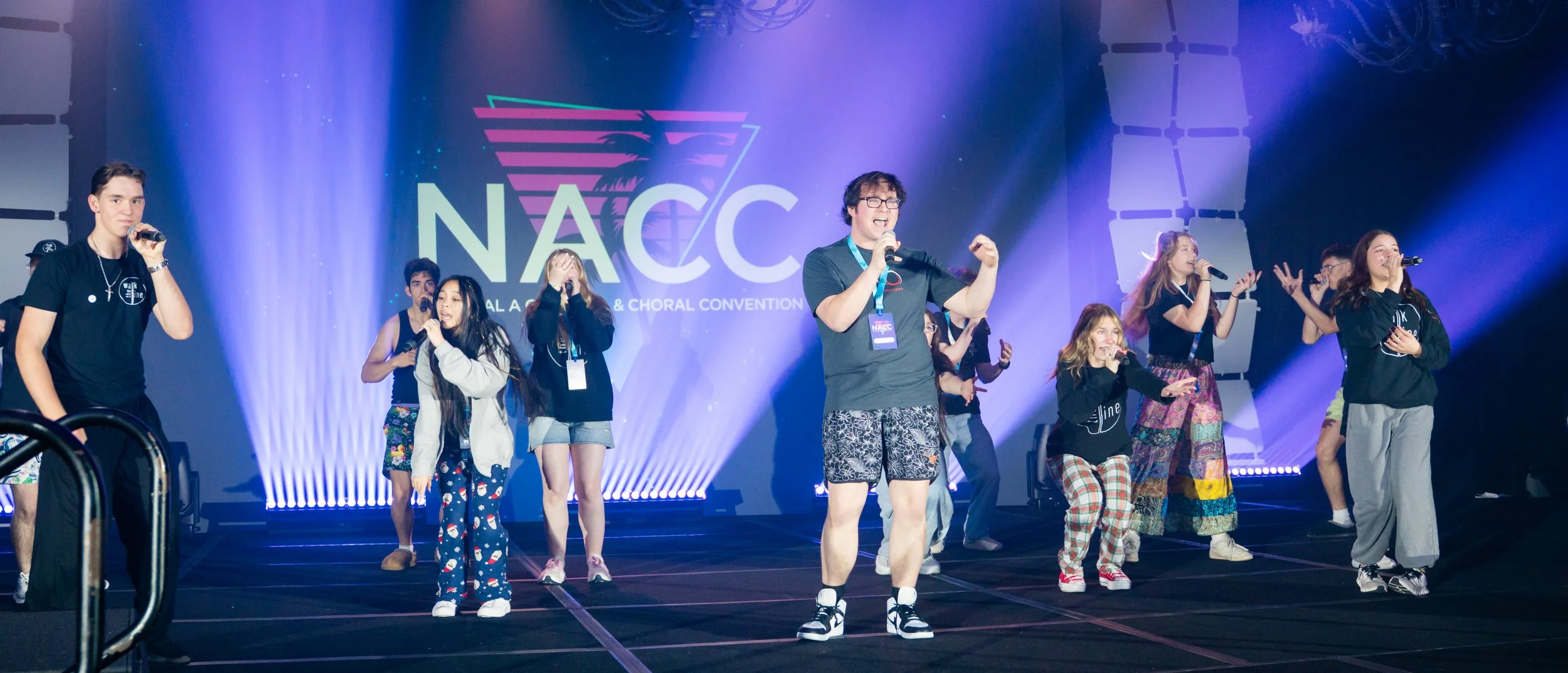 A group of kids and teens singing and performing on stage at a conference called NACAC, with a large screen behind them displaying the event's logo.