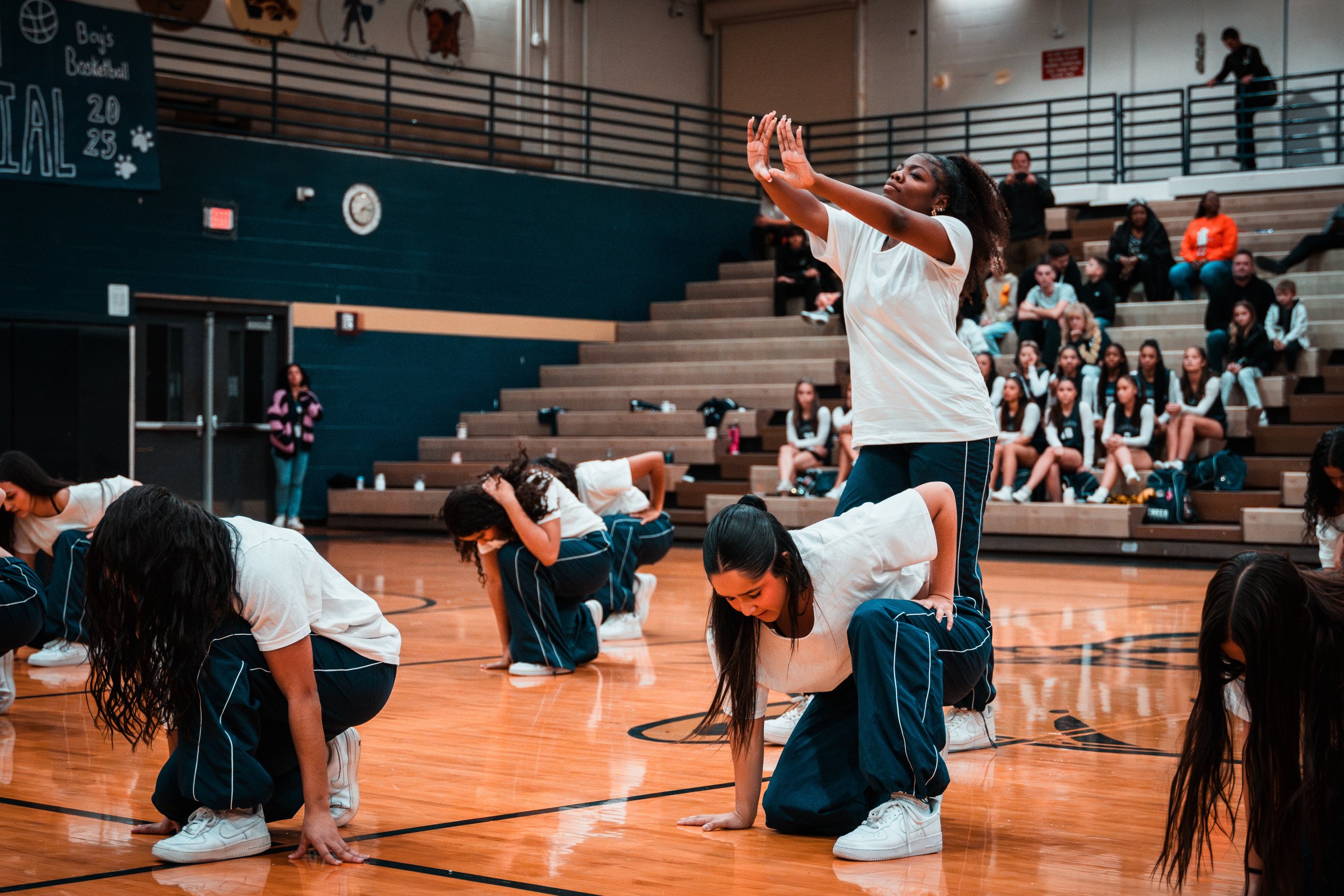 A group of students performing a cheerleading routine inside a gymnasium, with one girl standing and clapping her hands while others are down on their hands and knees.