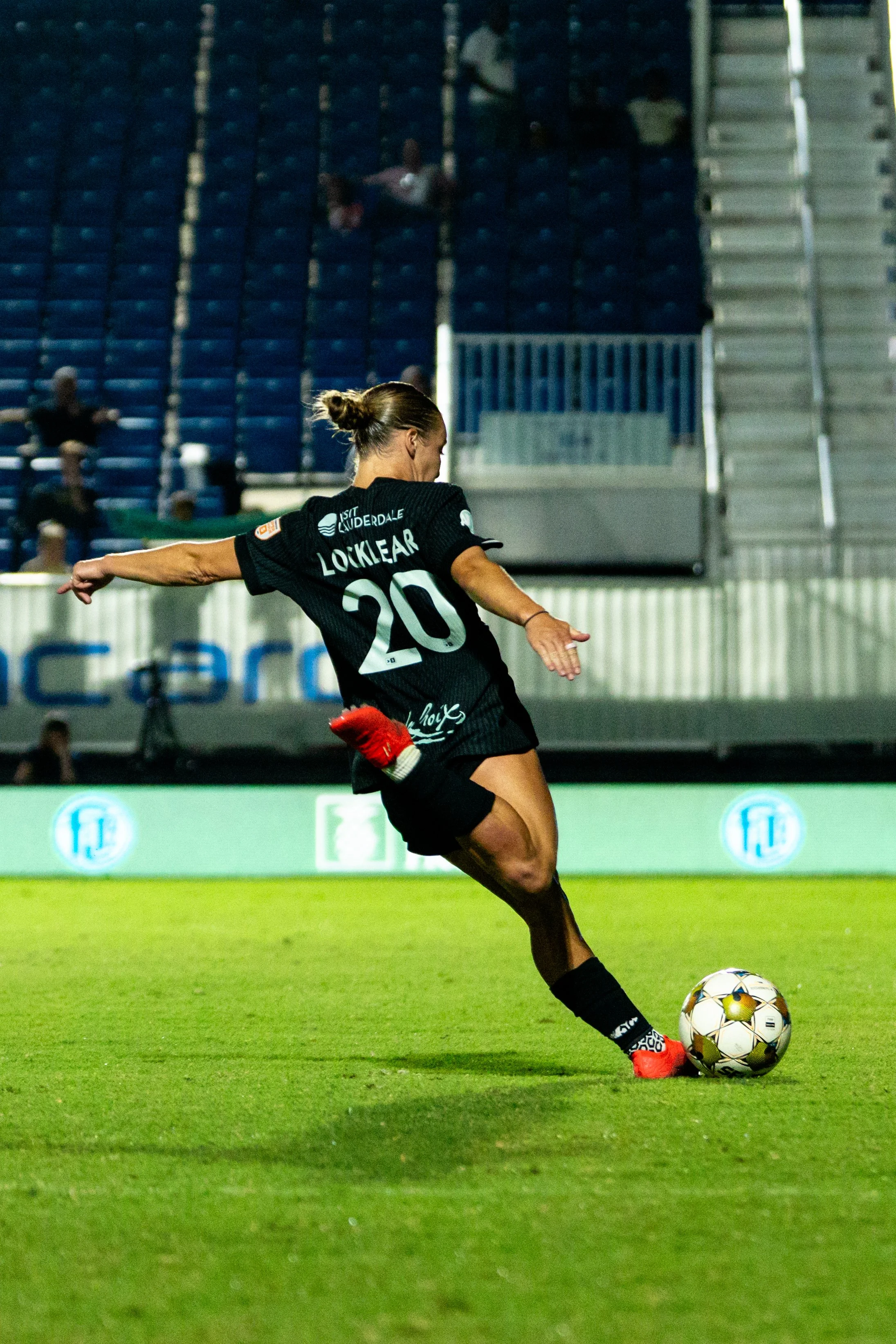 A female soccer player kicking a football on the field, wearing a black jersey with the number 20 and the name Locklear, with stadium seats in the background.