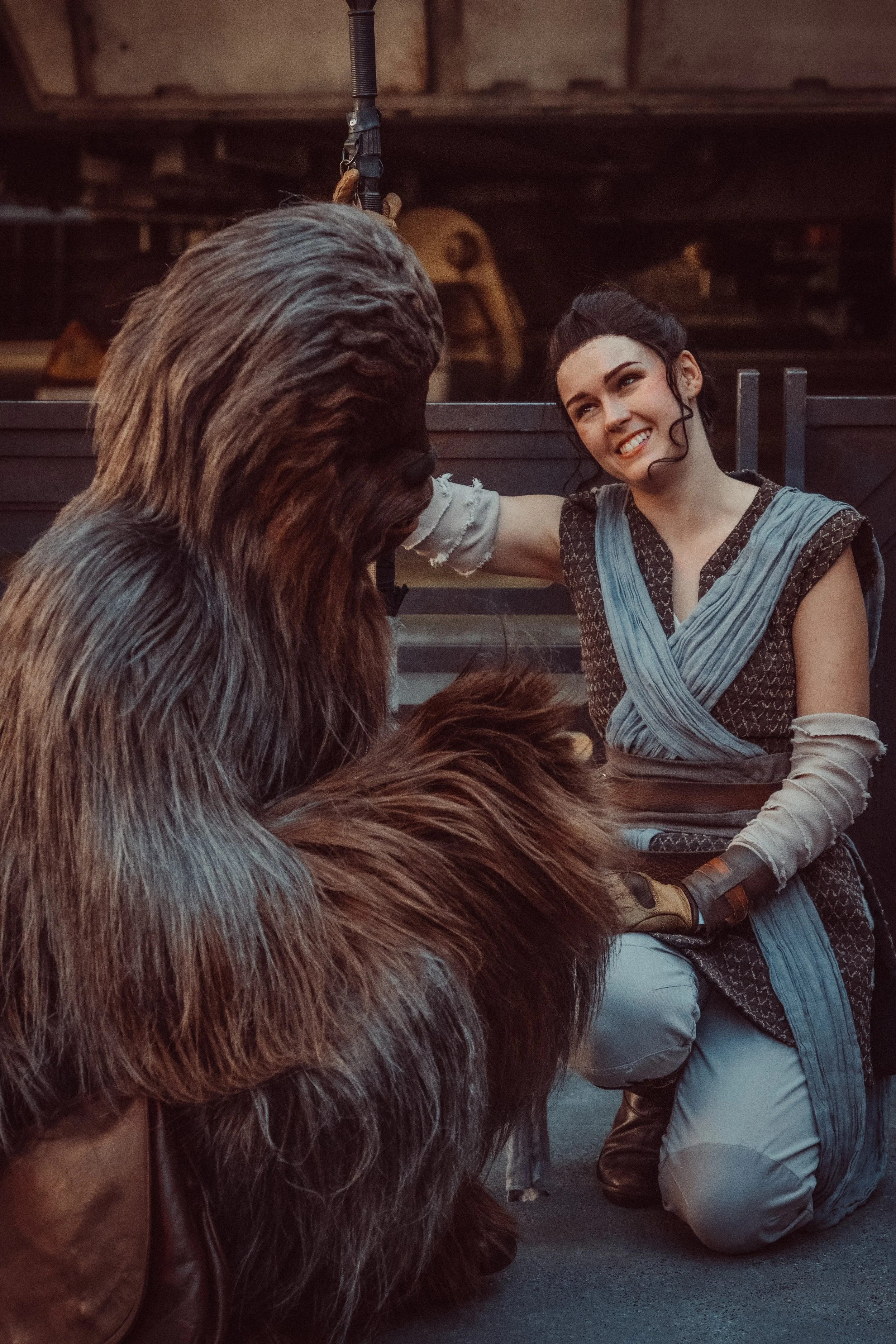 Woman with dark hair smiling and kneeling beside a person in a Chewbacca costume, in a shed or workshop setting.