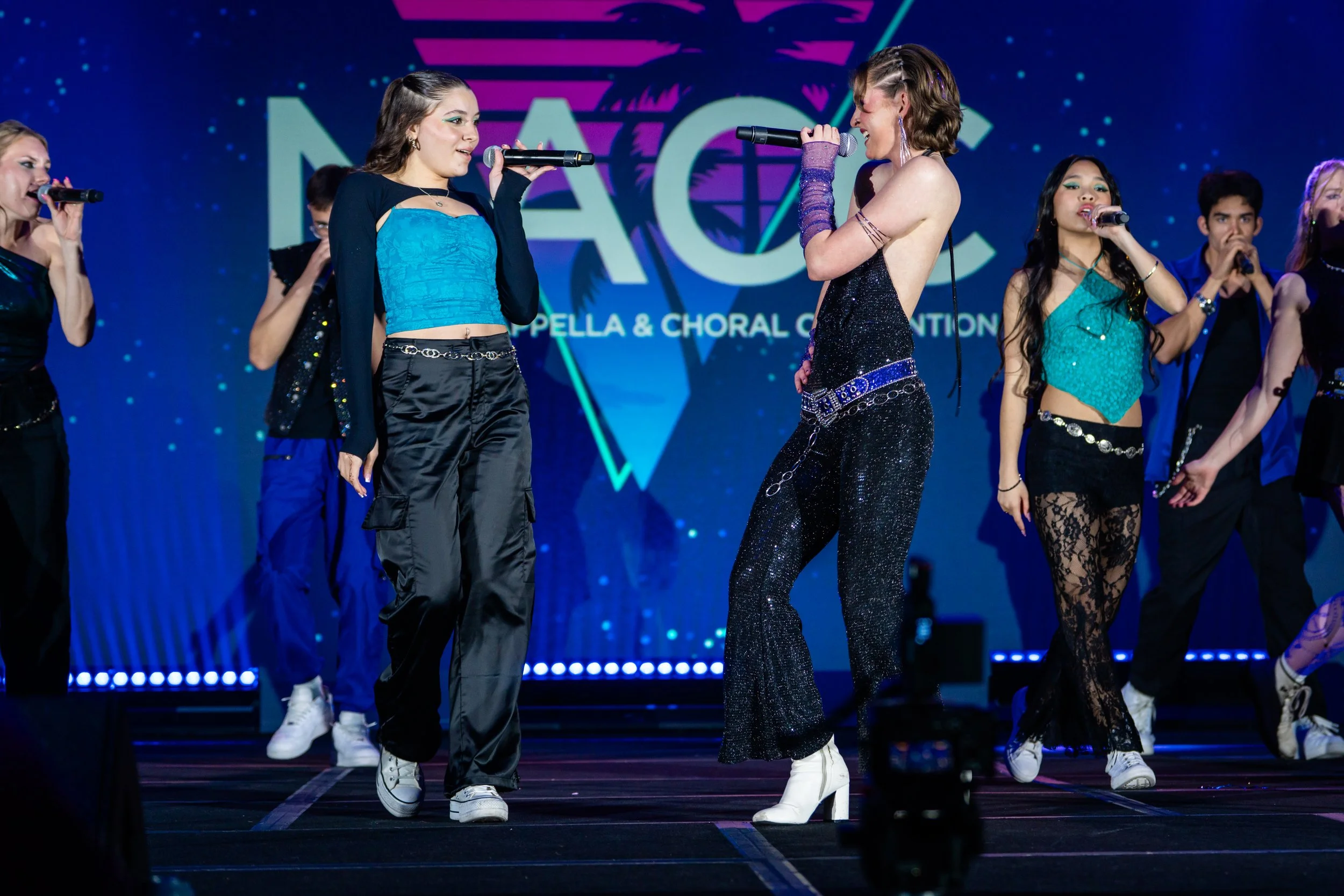 Young women and men performing on stage at a musical or singing event, dressed in colorful outfits, some holding microphones, with a backdrop displaying a logo and colorful lights.