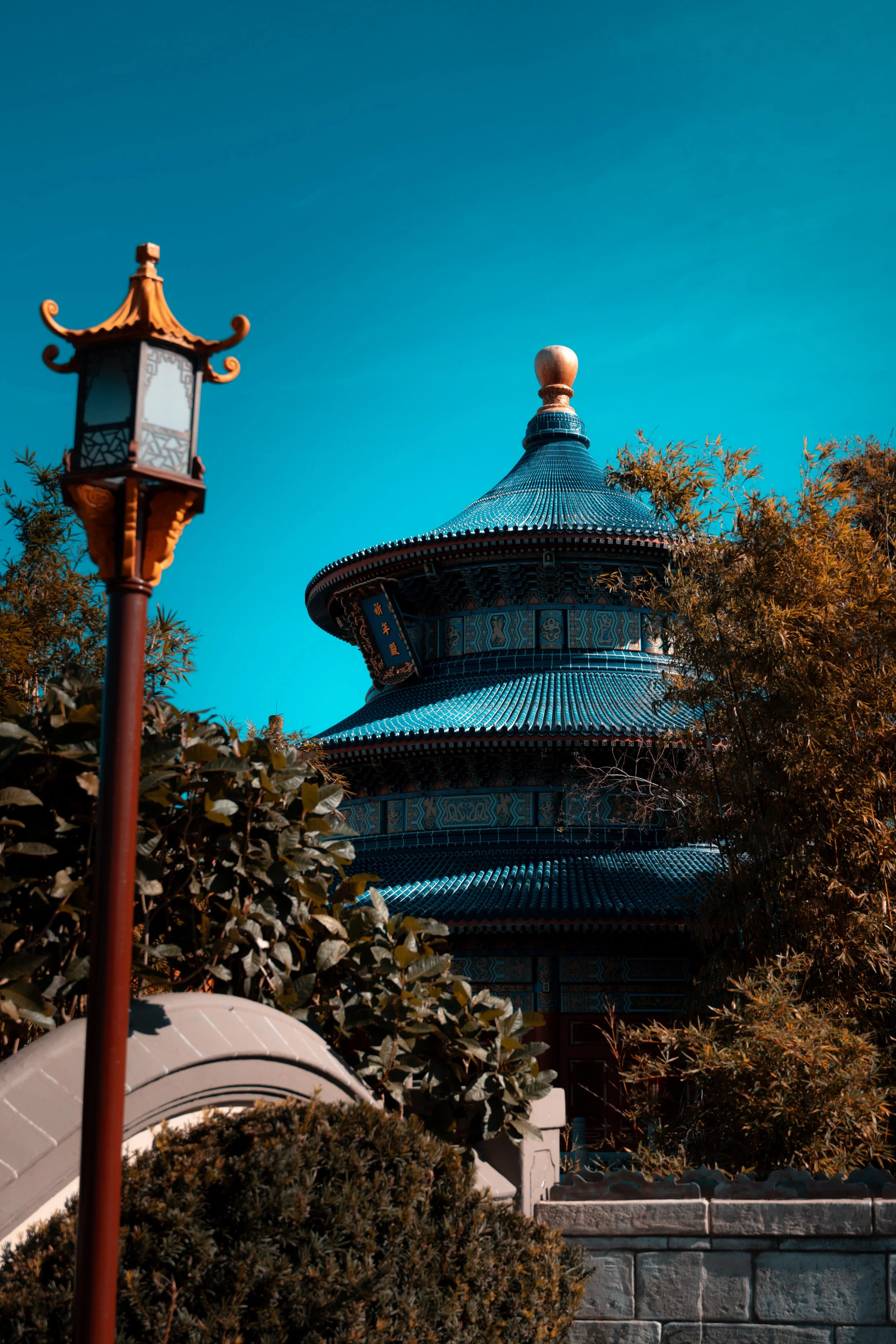 Traditional Asian pagoda-style building with ornate roof, surrounded by bushes and trees, with a decorative street lamp in the foreground and a clear blue sky in the background.