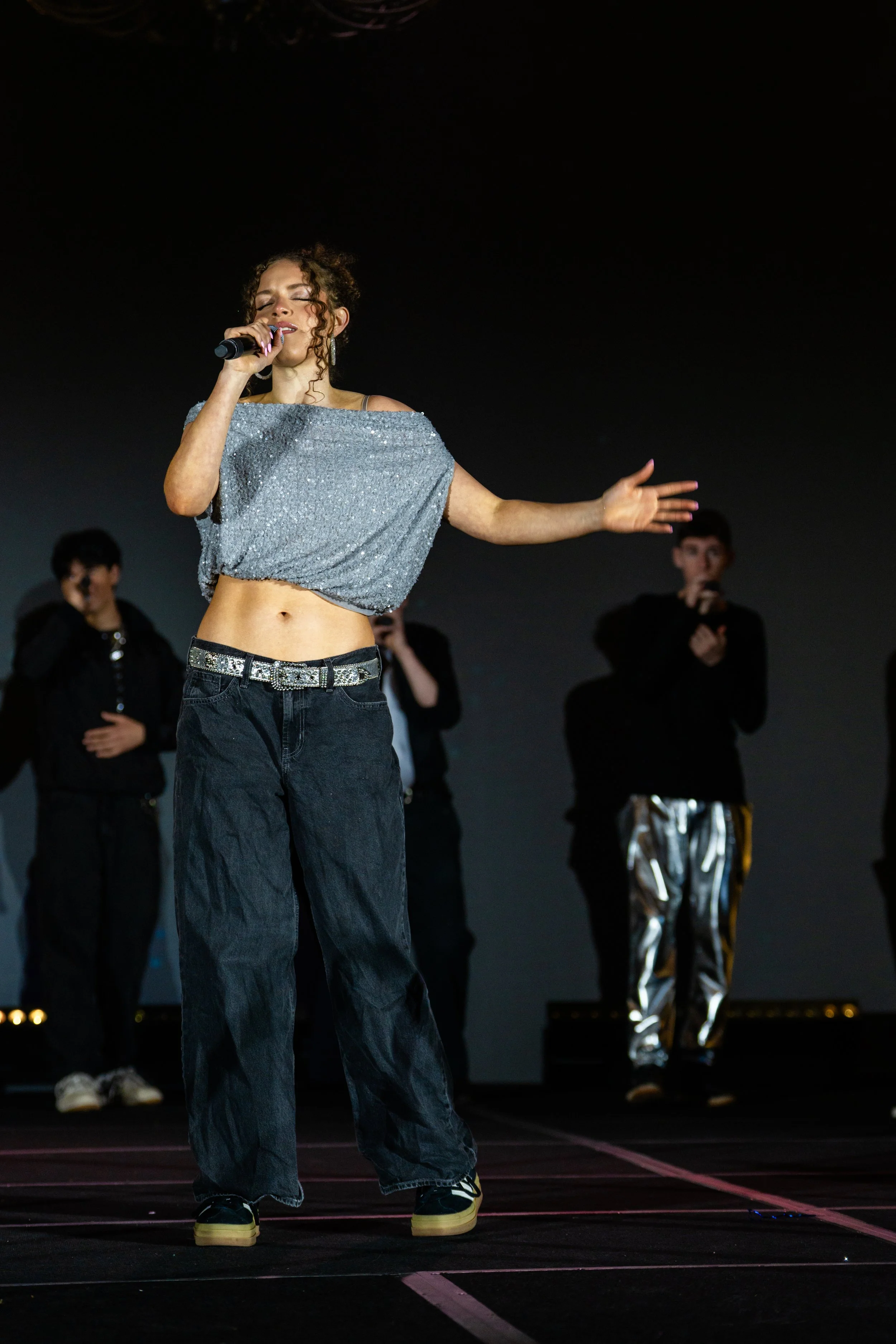 A woman singing into a microphone on stage, wearing a sparkly silver crop top and baggy black jeans, with three men in dark clothing standing behind her.