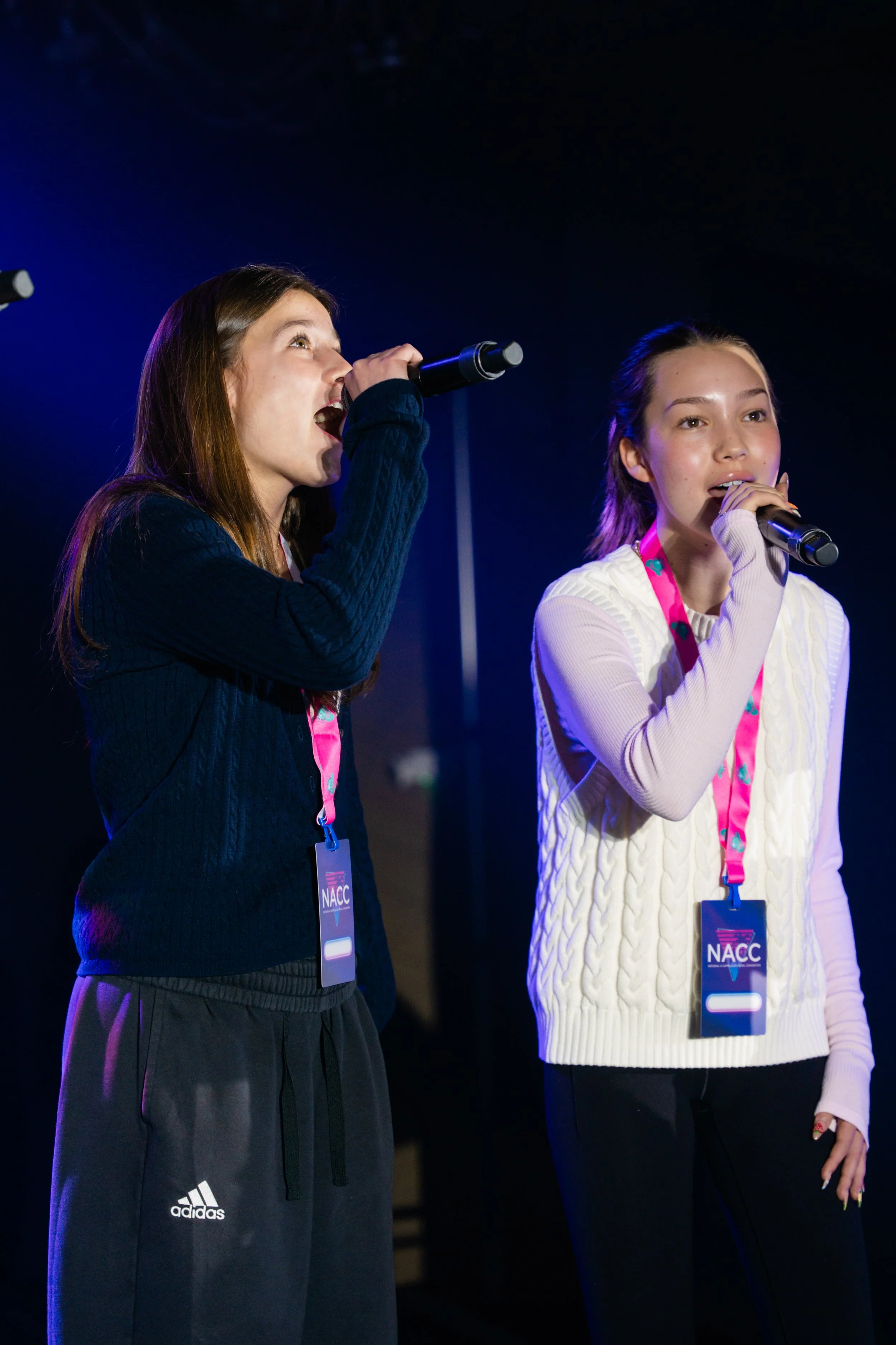 Two young women with microphones speaking at an event, wearing event badges with "NACC" on them, under dark lighting.