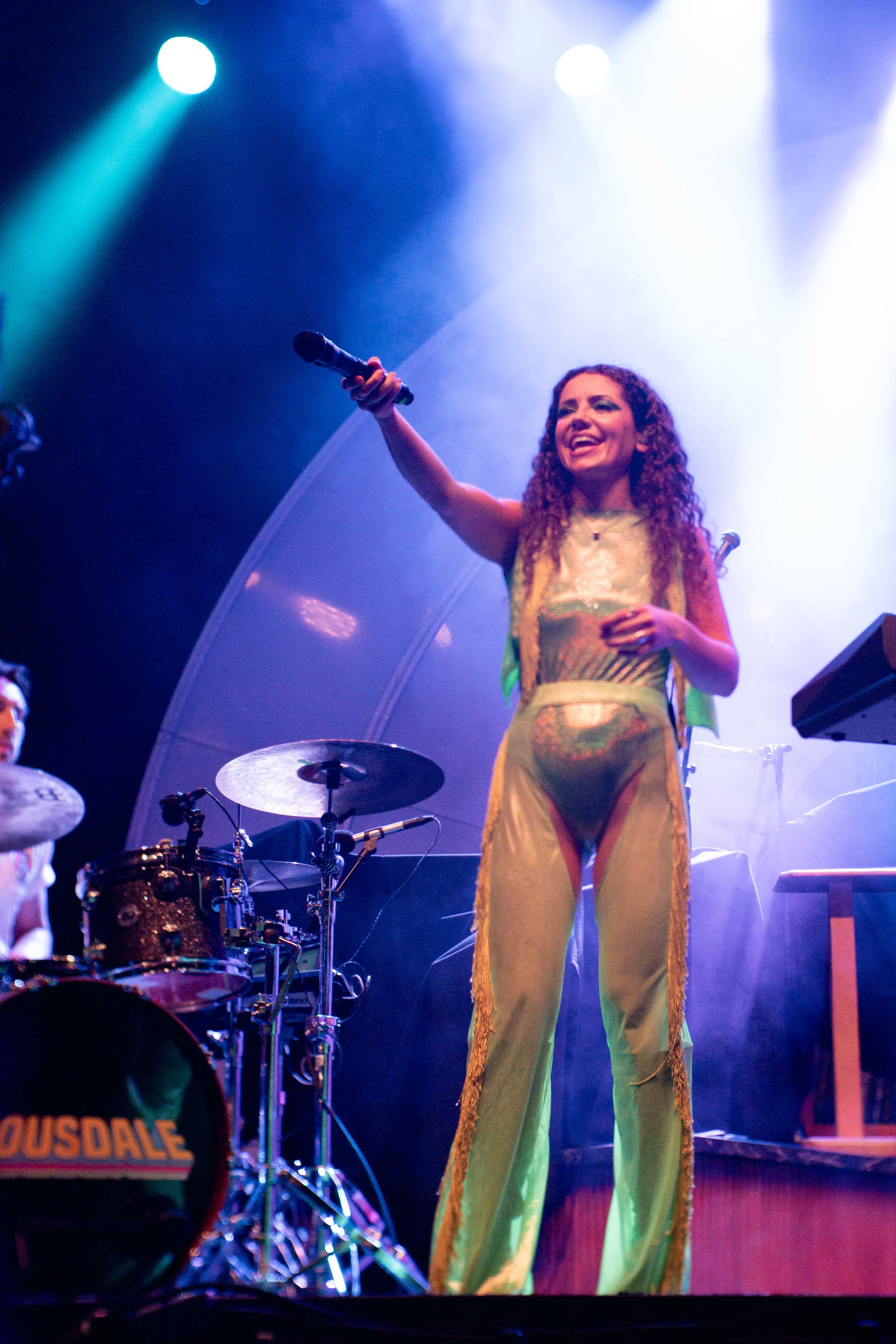 Performer on stage holding a microphone, smiling, with a drum set in the foreground and stage lights shining down.