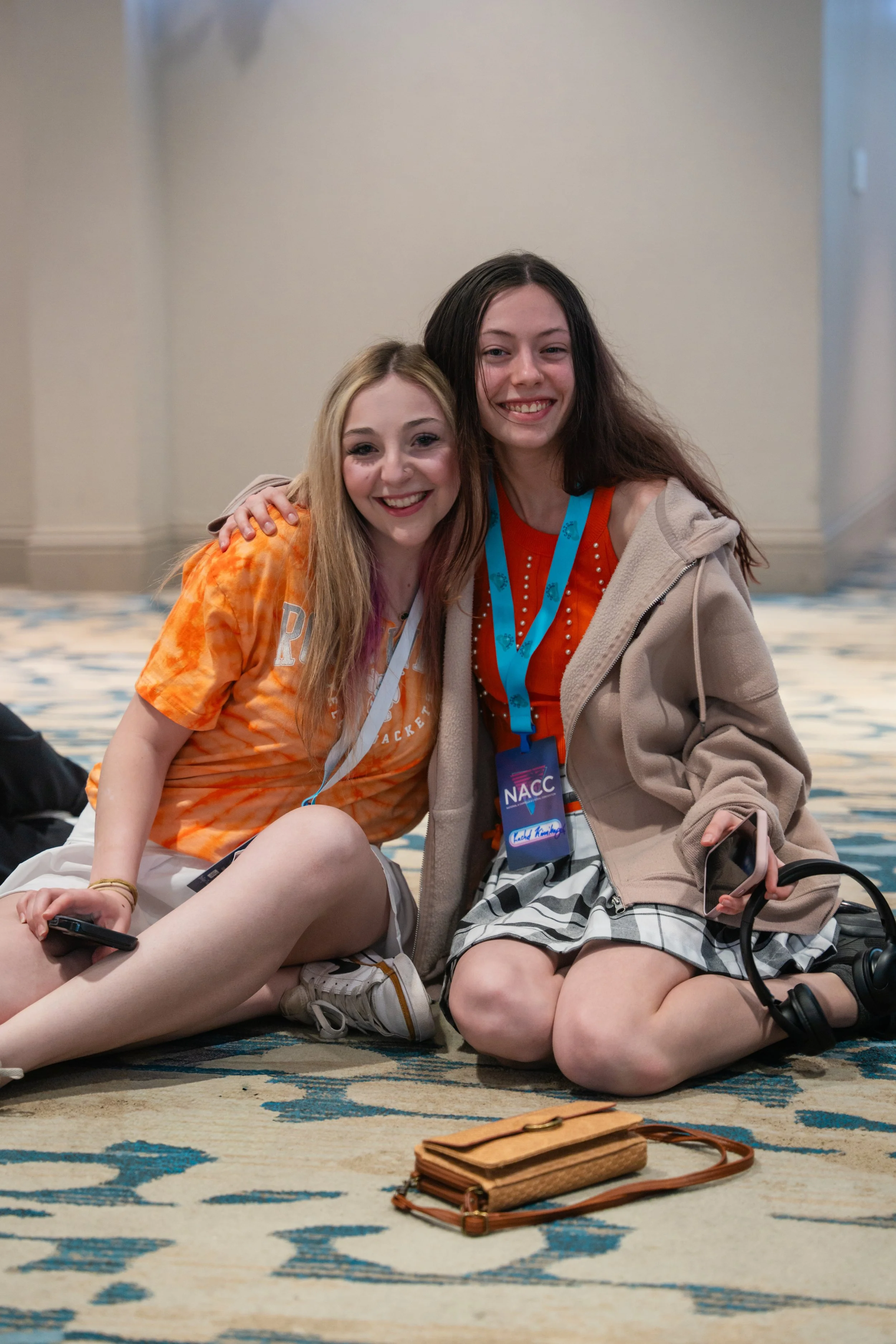 Two young women sitting on a patterned carpet at a conference, smiling at the camera. One is wearing an orange tie-dye shirt and the other a red shirt with a beige hoodie. They have conference badges around their necks. There are personal belongings 