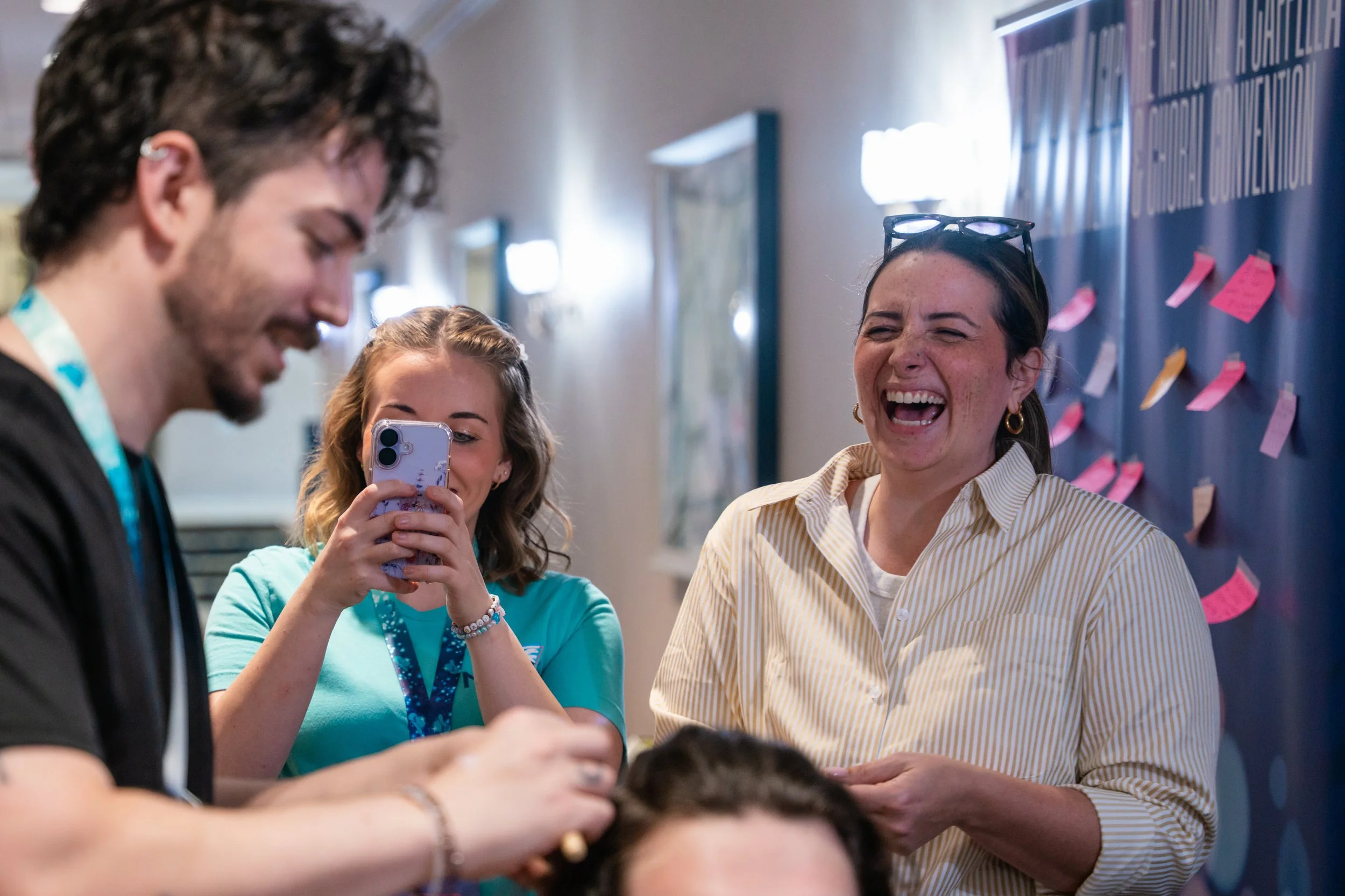 Four people are gathered in a hallway, with a woman laughing with her mouth open and sunglasses on her head. One woman is taking a selfie, and a man is on the left side with his face partially visible. The background features a board with pink and wh