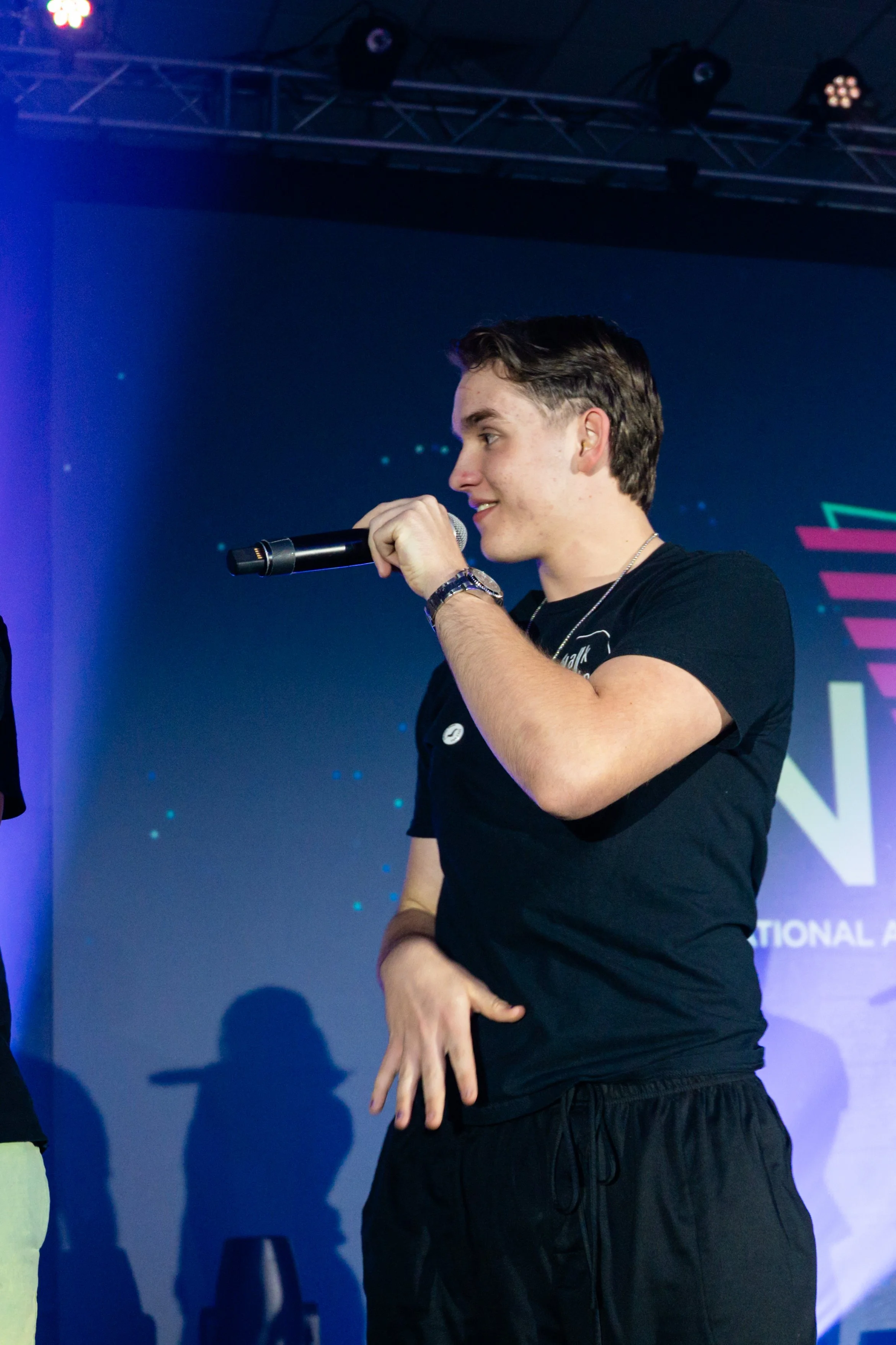 Young man holding a microphone on stage, wearing a black T-shirt and black pants, with background lights and a partial logo or text visible.