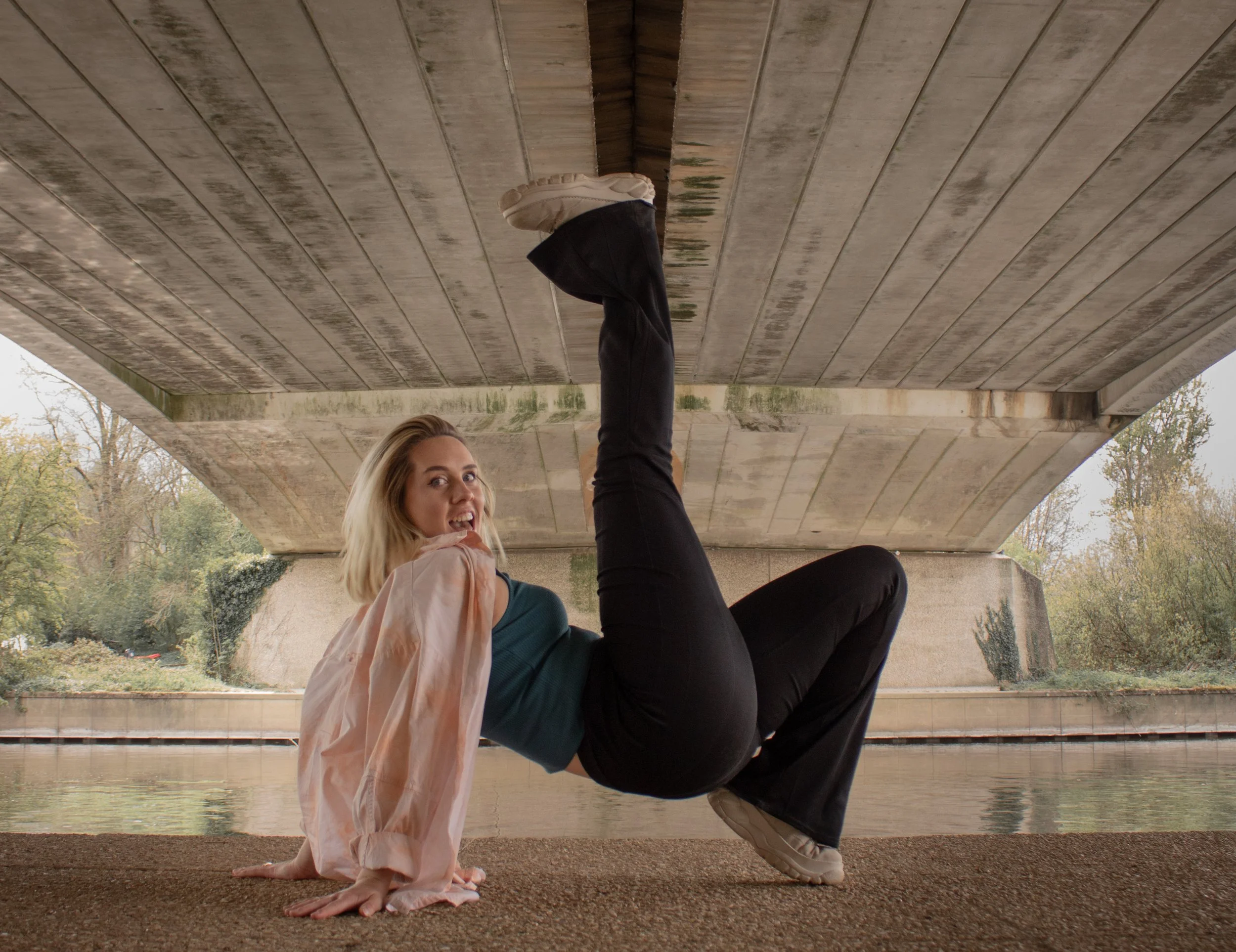 A woman in black pants and a teal top, with a light-colored jacket, is posing under a bridge with her hands and dominant leg on the ground and her other leg raised against the bridge support, looking at the camera with her tongue slightly out. The scene is outdoors near a body of water with trees in the background.