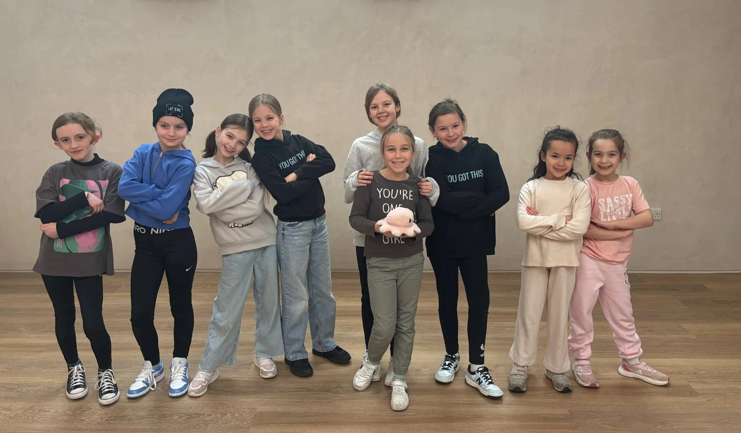 Group of nine young girls standing indoors on a hardwood floor, smiling at the camera, with one girl holding a plush toy, wearing casual and athletic clothing.