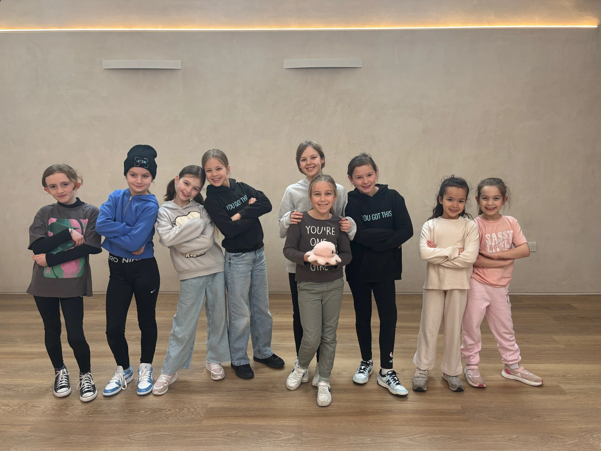 Group of nine young girls standing together in a room with wooden floors and plain walls, smiling and posing for the camera.