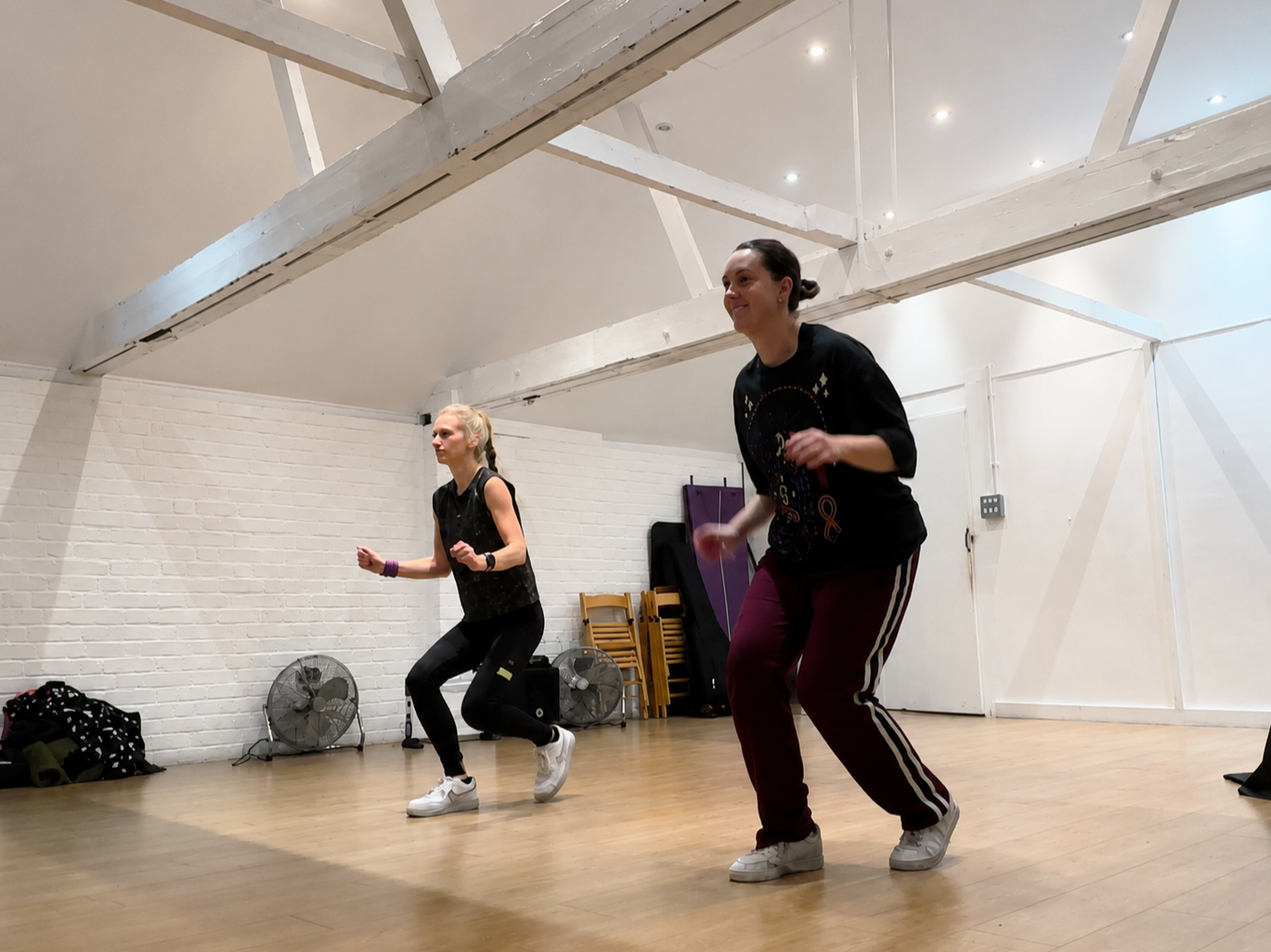 Two women dancing in a studio with white brick walls, wooden floor, and ceiling beams. There are fans, chairs, and bags in the background.