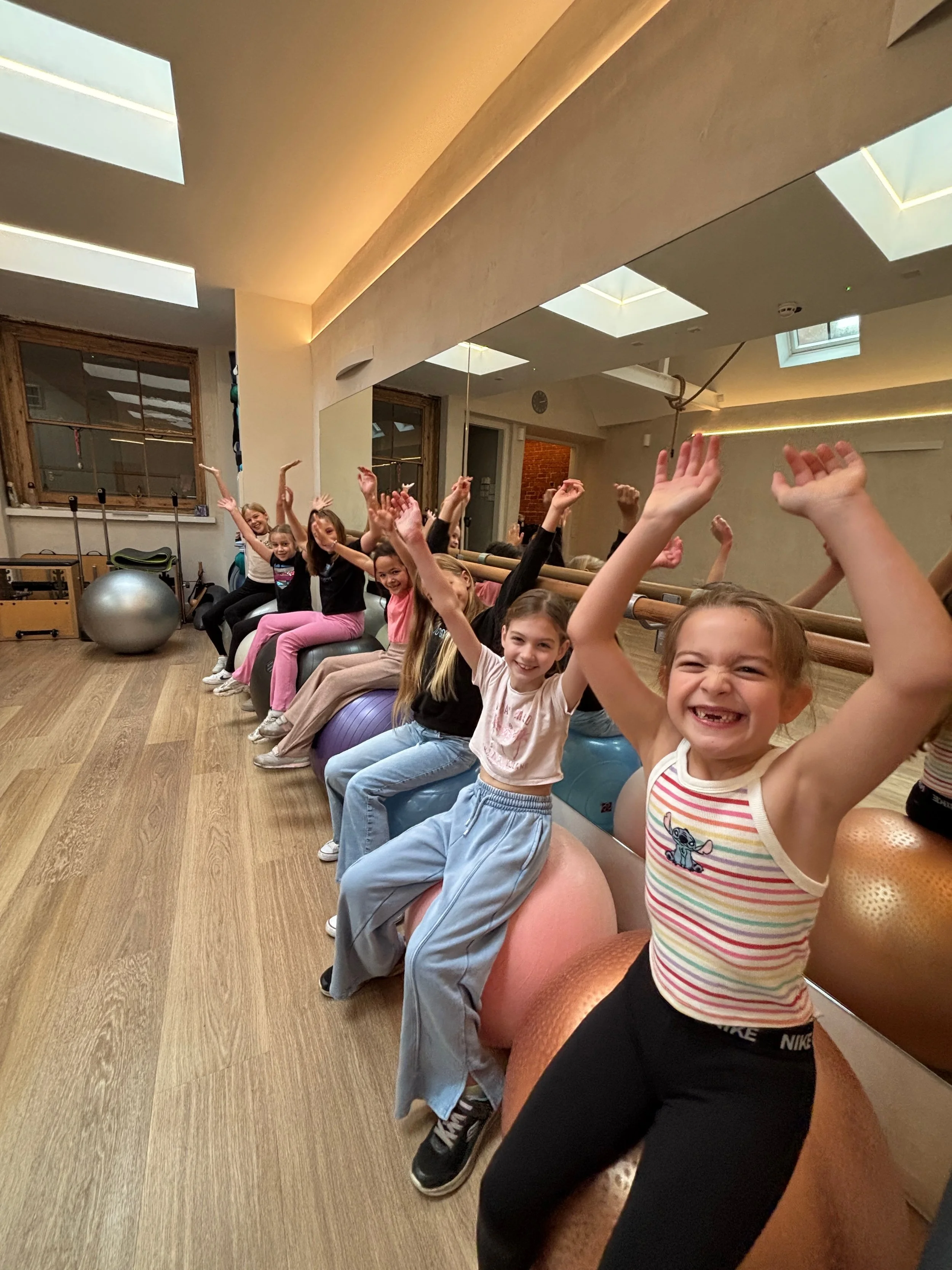 A group of children sitting on exercise balls in a fitness studio, smiling and raising their hands in the air.