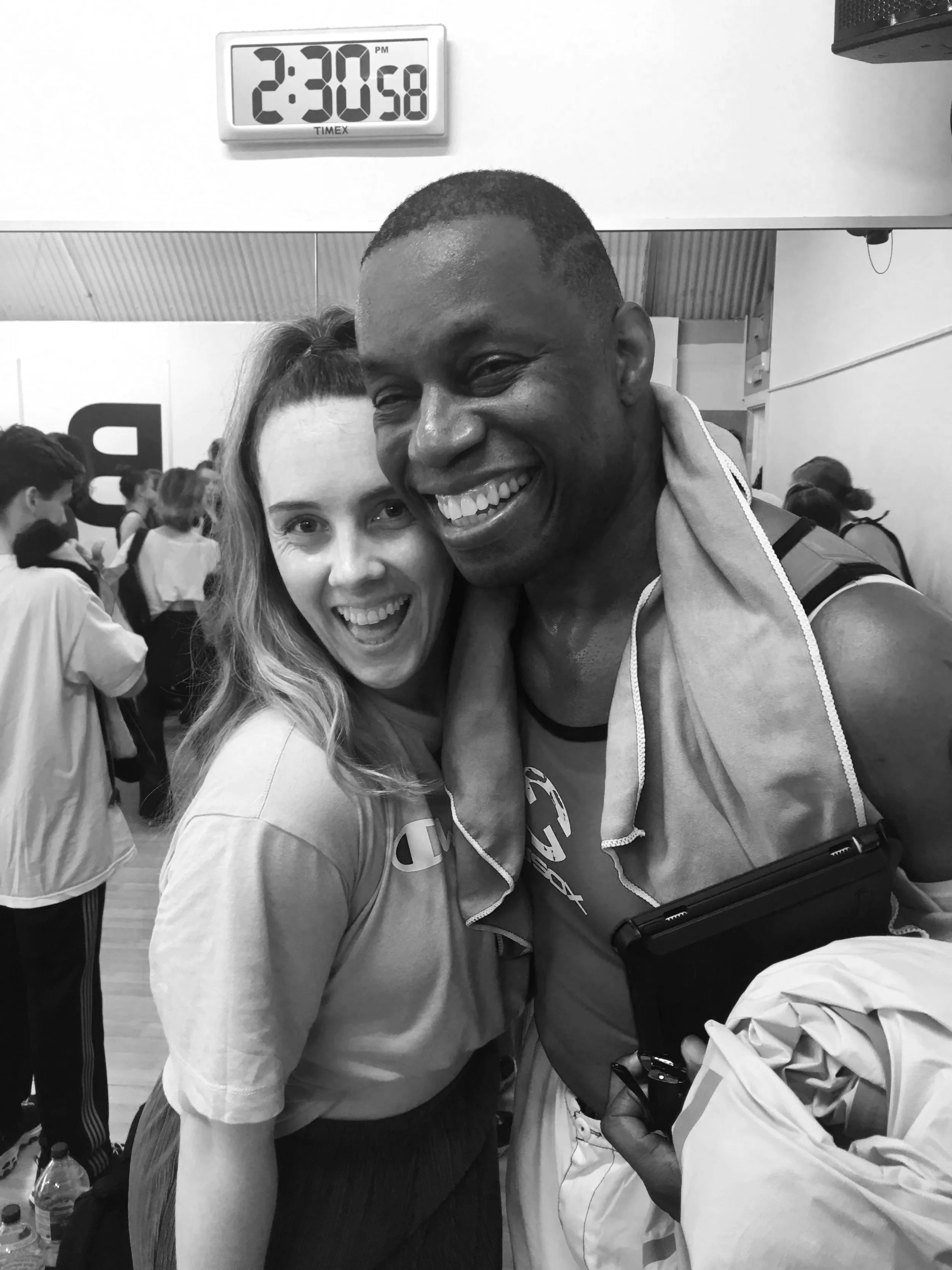 A smiling woman and man hugging each other at a sports event, with a crowd of people in the background. The man is holding a small device and a jacket, and they are in an Olympic venue with a large clock showing 2:30.