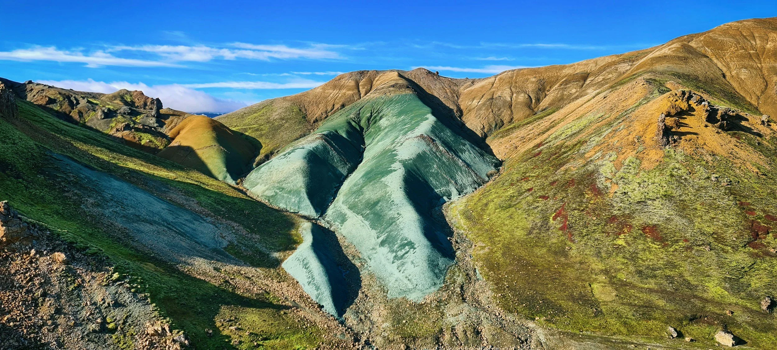 Colorful mountains with green, brown, and gray slopes under a blue sky with wispy clouds.