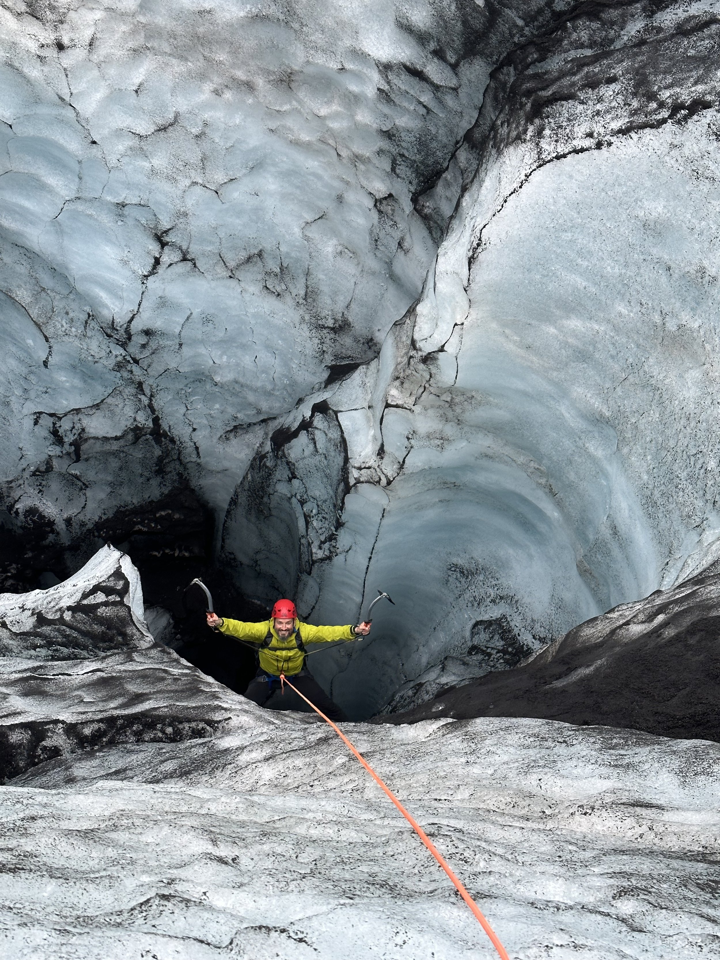 A person in a yellow jacket and red helmet descenting a glacier using ice axes and a rope in a crevasse with ice formations.