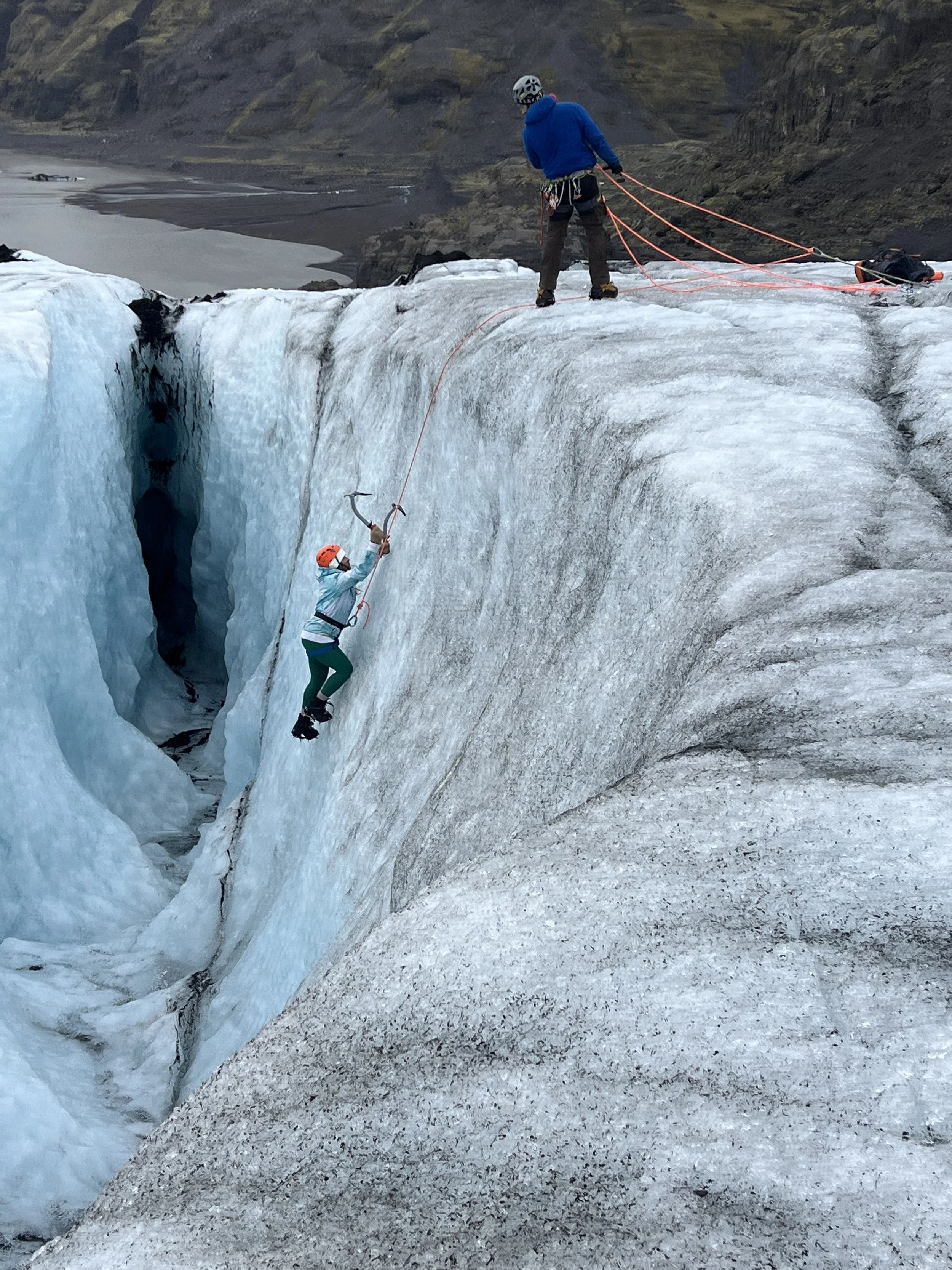 Two people, dressed in outdoor gear and helmets, are ice climbing on a glacier. One climber is mid-ascent, while the other manages the ropes at the top. The glacier has icy, rugged surfaces, with a snow-covered mountain landscape in the background.
