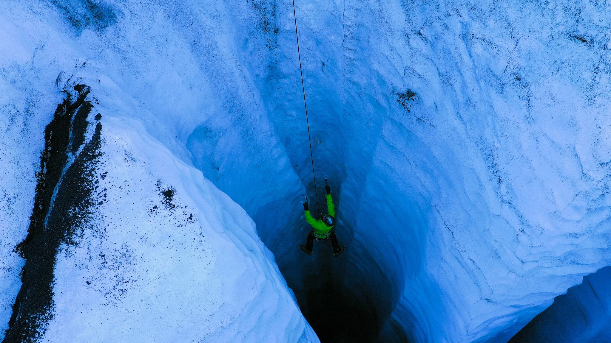 A person wearing a green jacket and black helmet is rappelling into a deep crevasse in a glacier with blue ice walls.