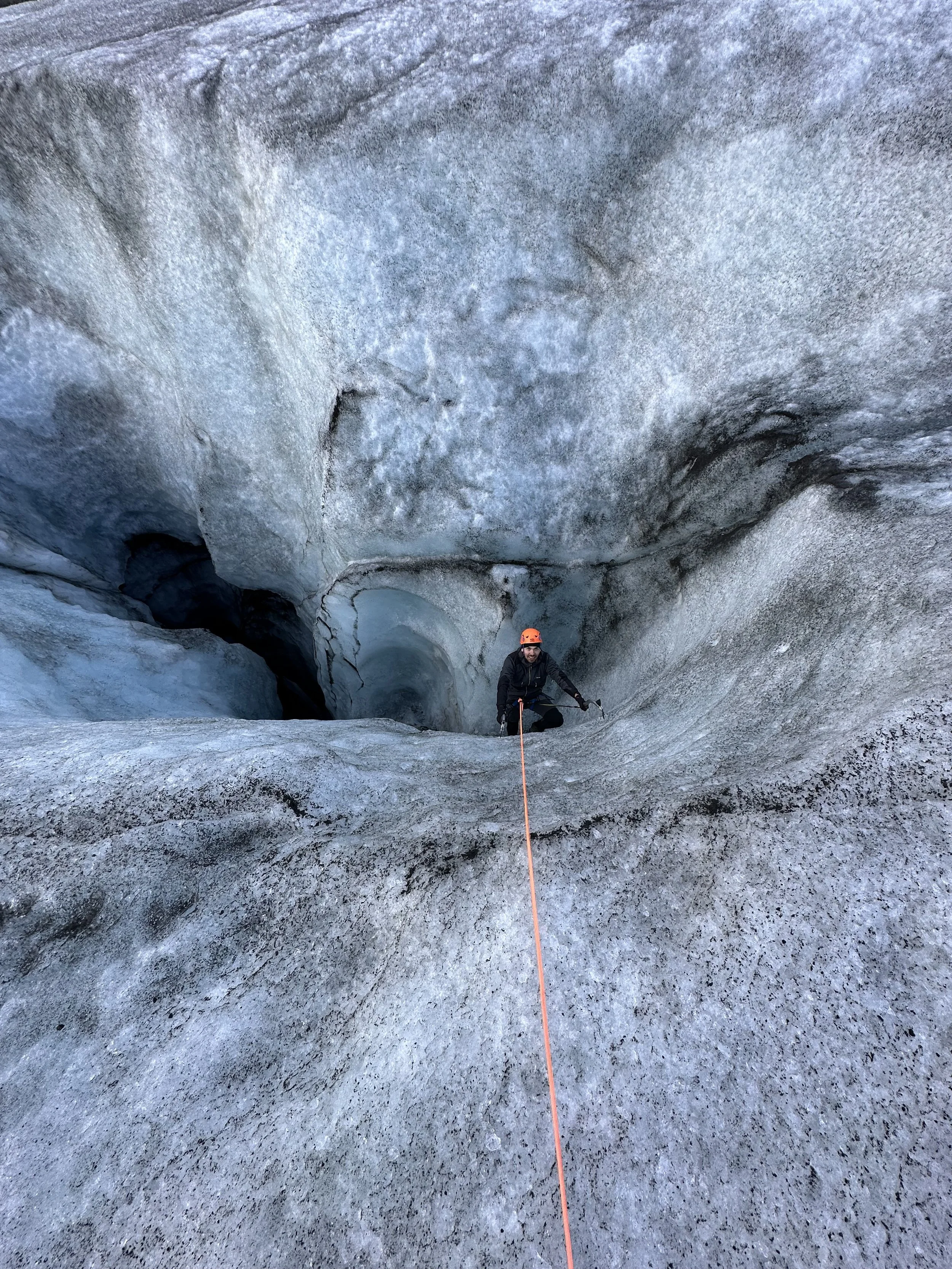 Individual wearing an orange helmet and black outdoor gear sitting on a glacier ice surface with cracks and crevices, holding an ice axe, with a rope extending toward the camera.