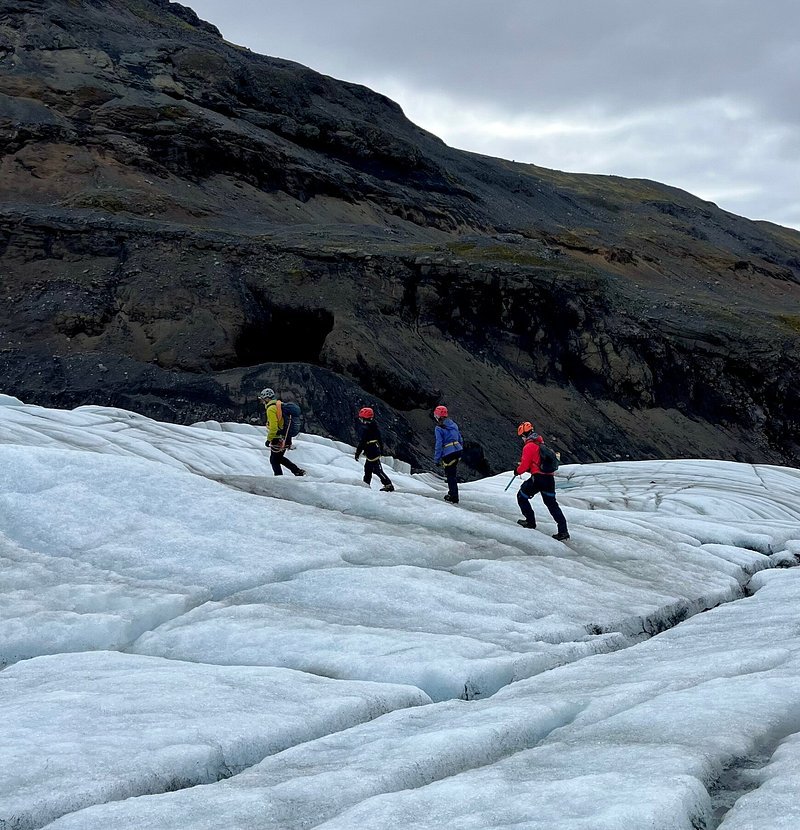 Four people wearing helmets and outdoor gear ice climbing or hiking on a glacier with rugged, dark mountains in the background under cloudy skies.