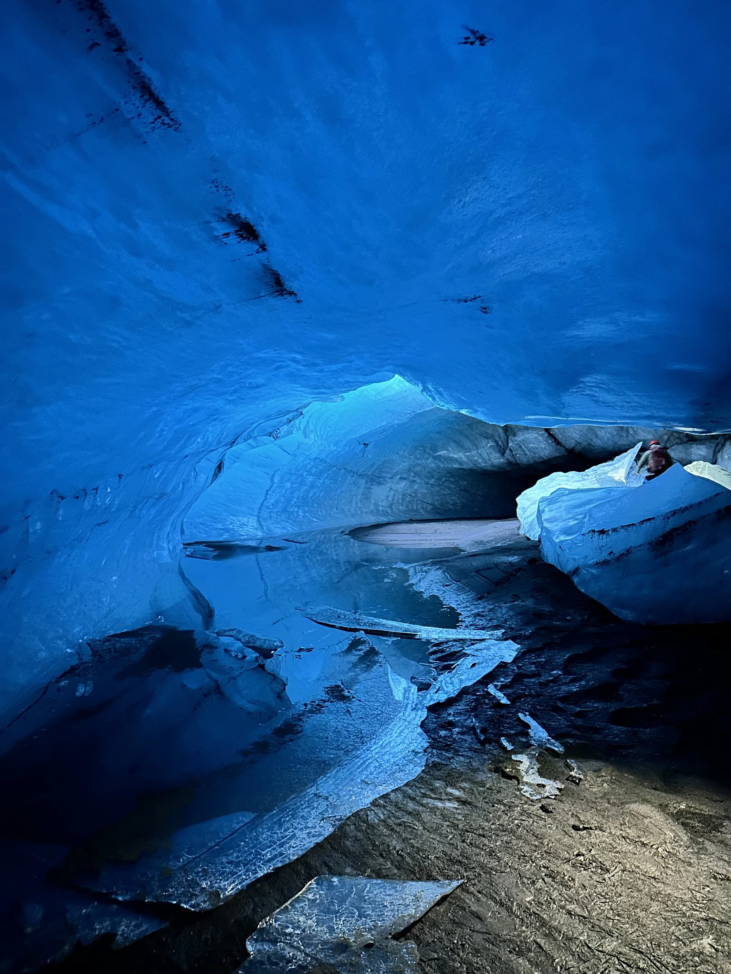 View inside a blue ice cave with icy blue walls and ceiling, and a muddy ground with patches of ice.