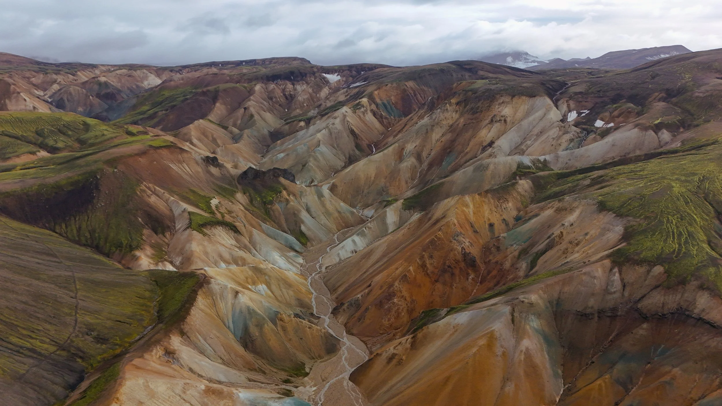 Aerial view of a mountainous region with colorful, layered terrain, small patches of snow, and a winding river valley.