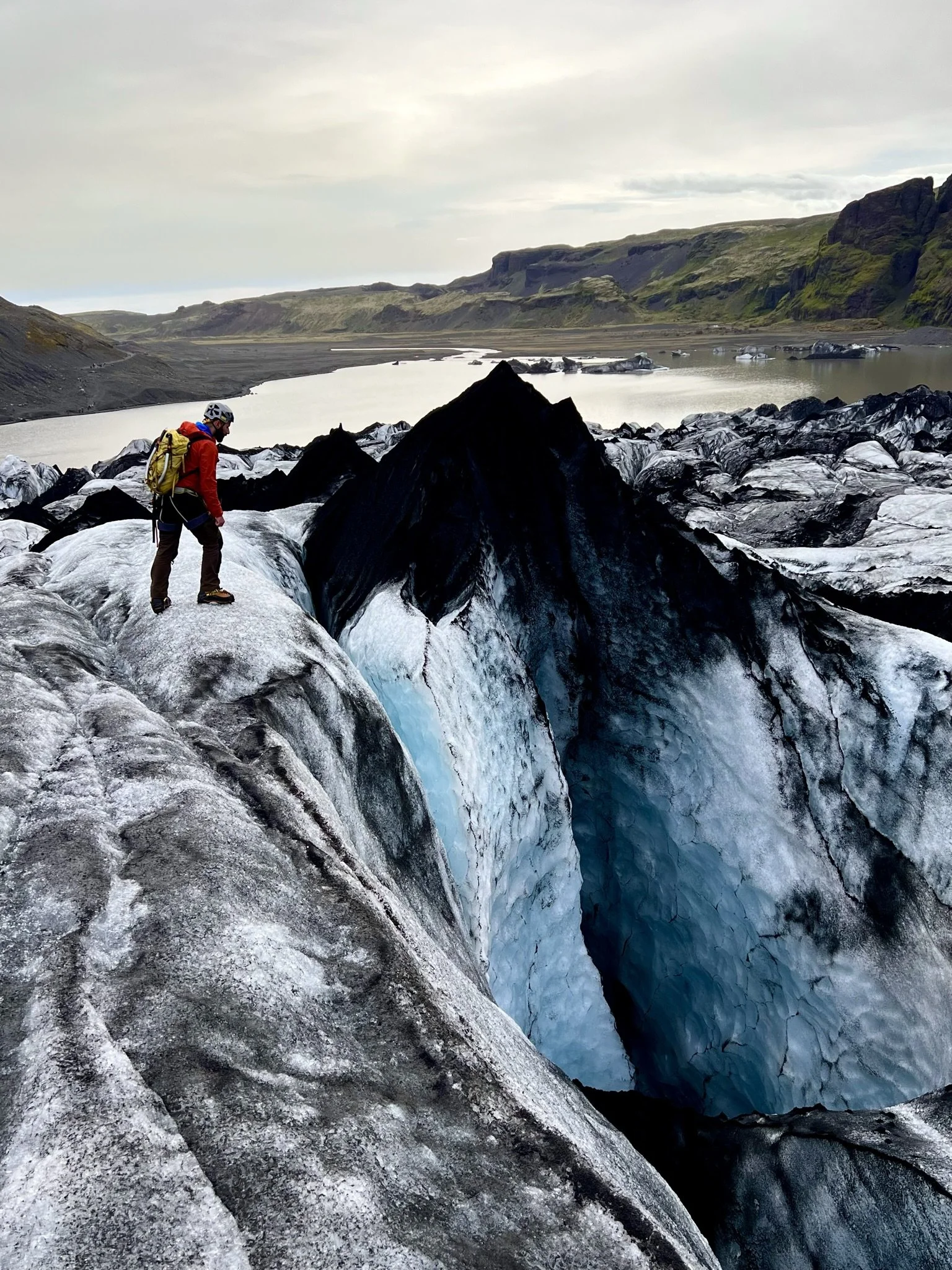 A person in outdoor gear standing on ice near a glacier with water and distant green hills in the background.