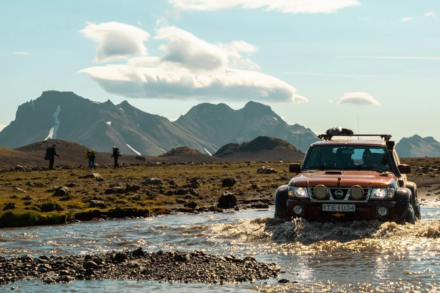 An off-road vehicle crossing a shallow river in a mountainous landscape with three hikers walking in the background, under a blue sky with scattered clouds.