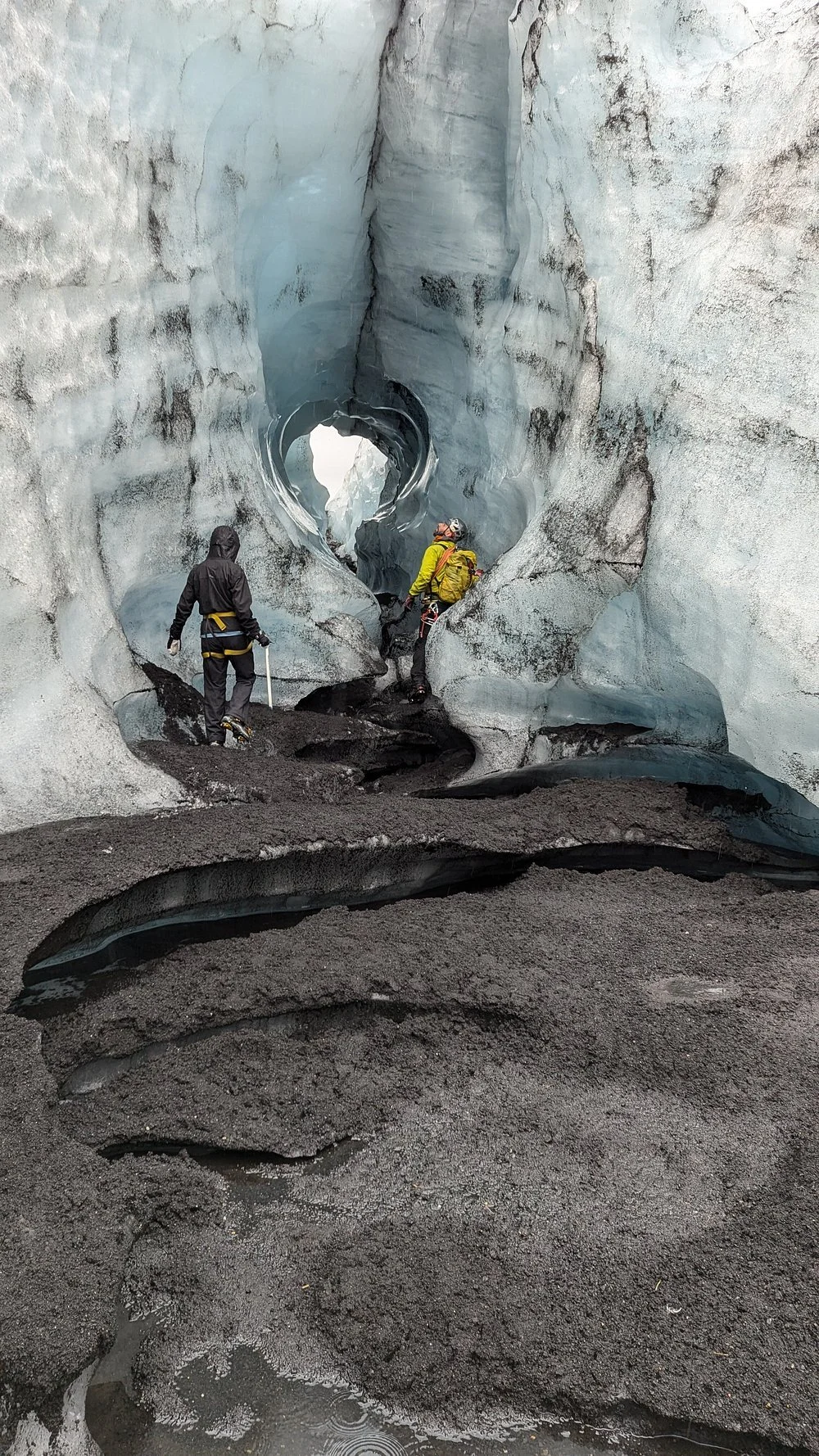 Two people in outdoor gear exploring a narrow icy crevasse with ice walls and dark volcanic sand on the floor.
