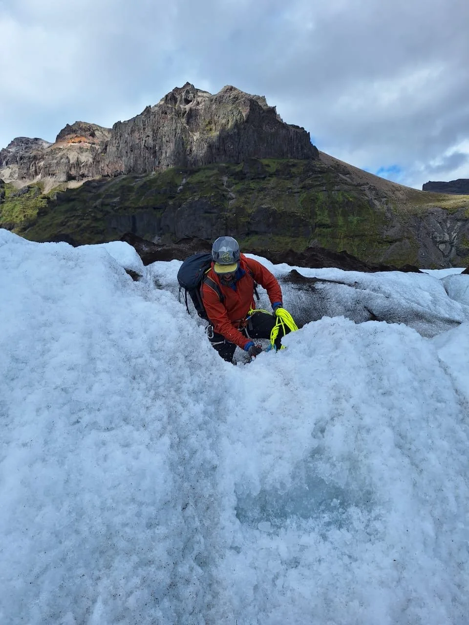 Mountaineer climbing snow-covered ice with a mountain and cloudy sky in the background.