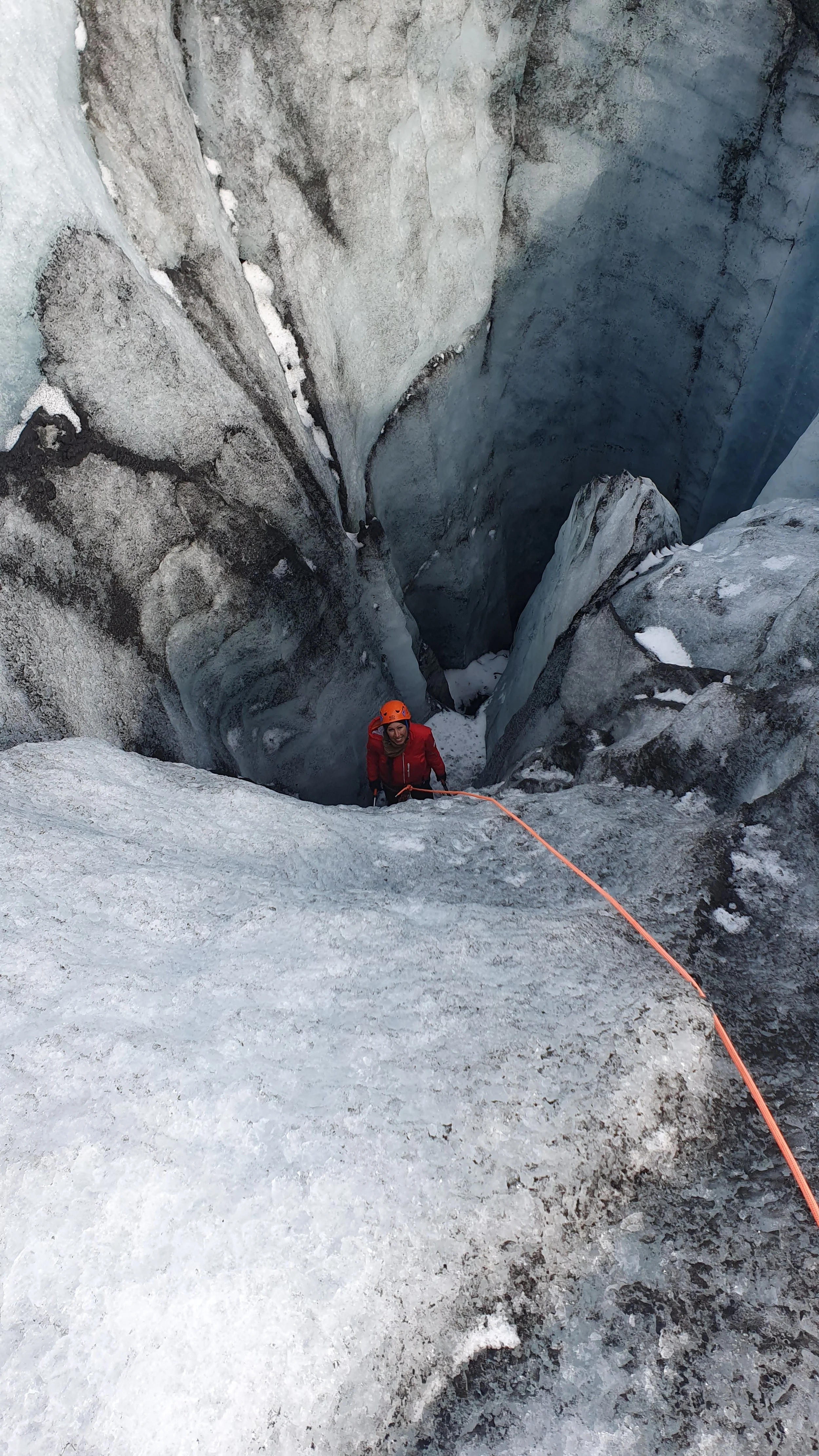 A person wearing a red jacket and an orange helmet ice climbing on a steep ice wall, with a safety rope attached to their harness.