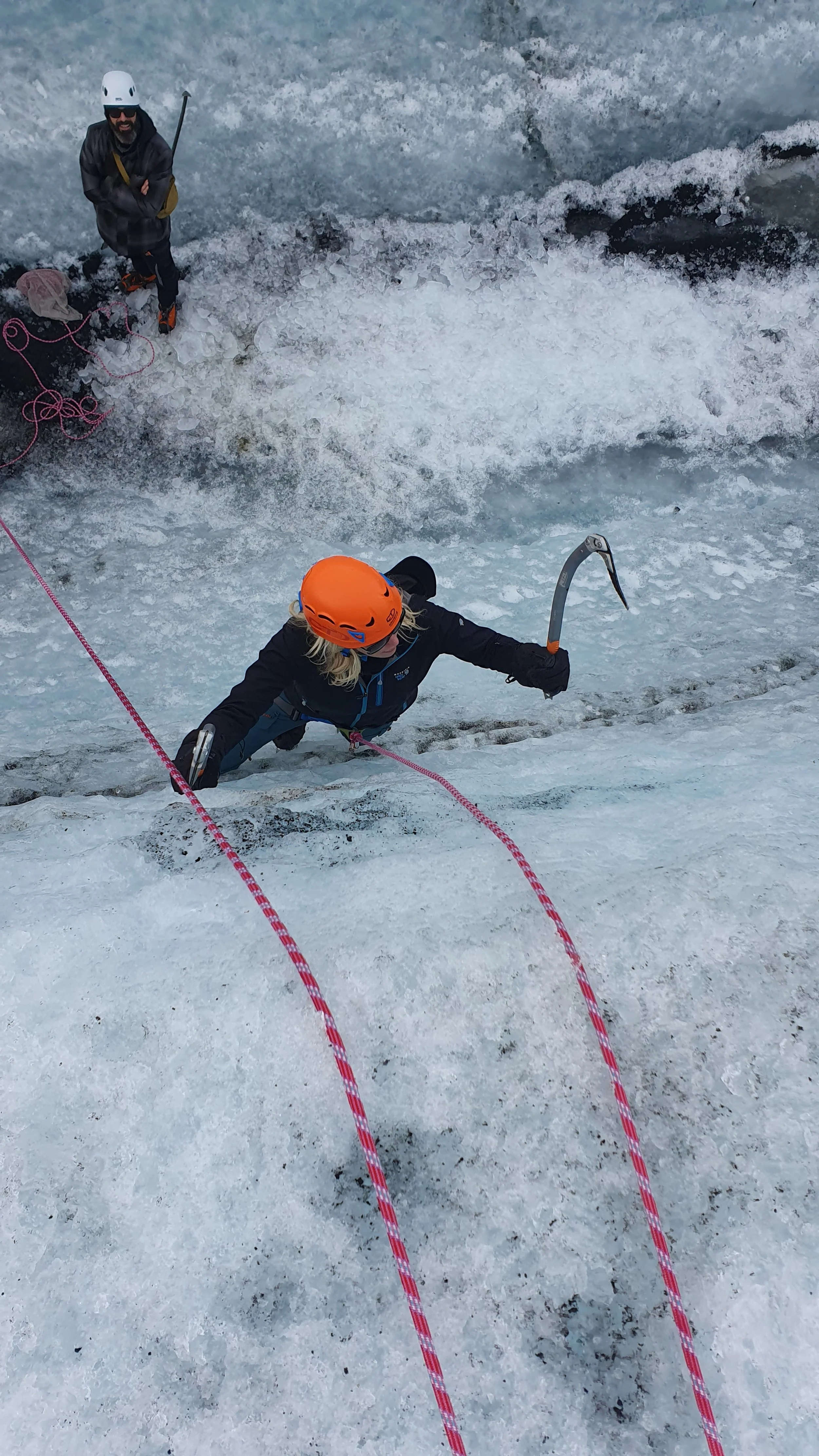 Two climbers ascending an icy rock face, one wearing an orange helmet and harness, the other standing on the ice wearing a white helmet, with climbing ropes and icy surroundings.