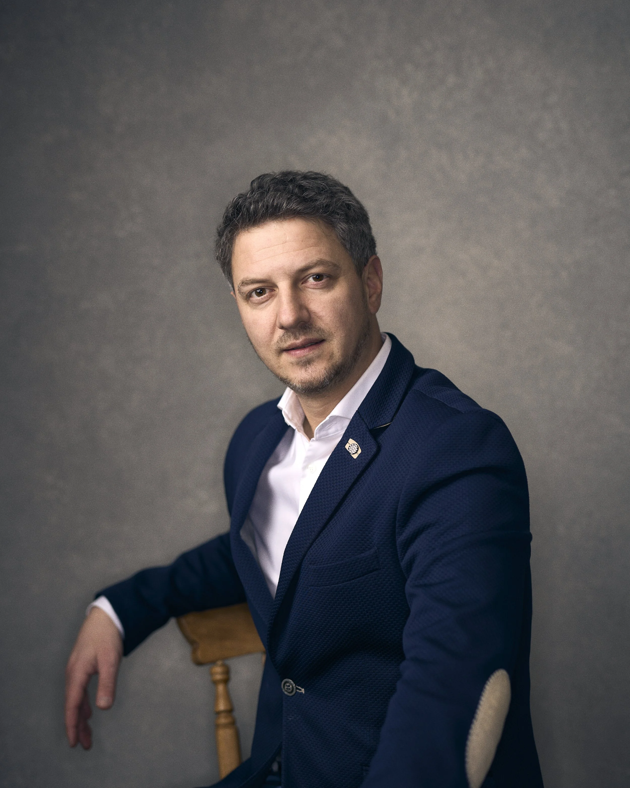 Portrait of a man with dark hair and light skin, wearing a navy blue blazer and white shirt, sitting on a wooden chair against a gray textured background.