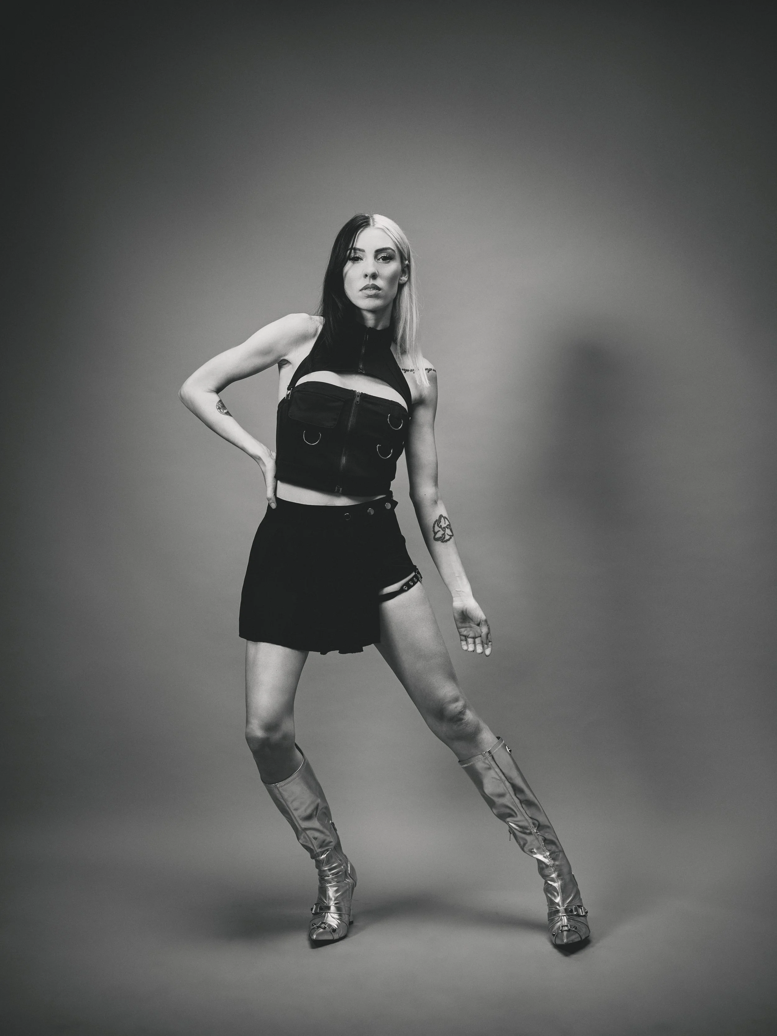 Black and white photo of a woman standing confidently with one hand on her hip and the other relaxed by her side, wearing a sleeveless top, a short skirt, and high boots, against a plain background.