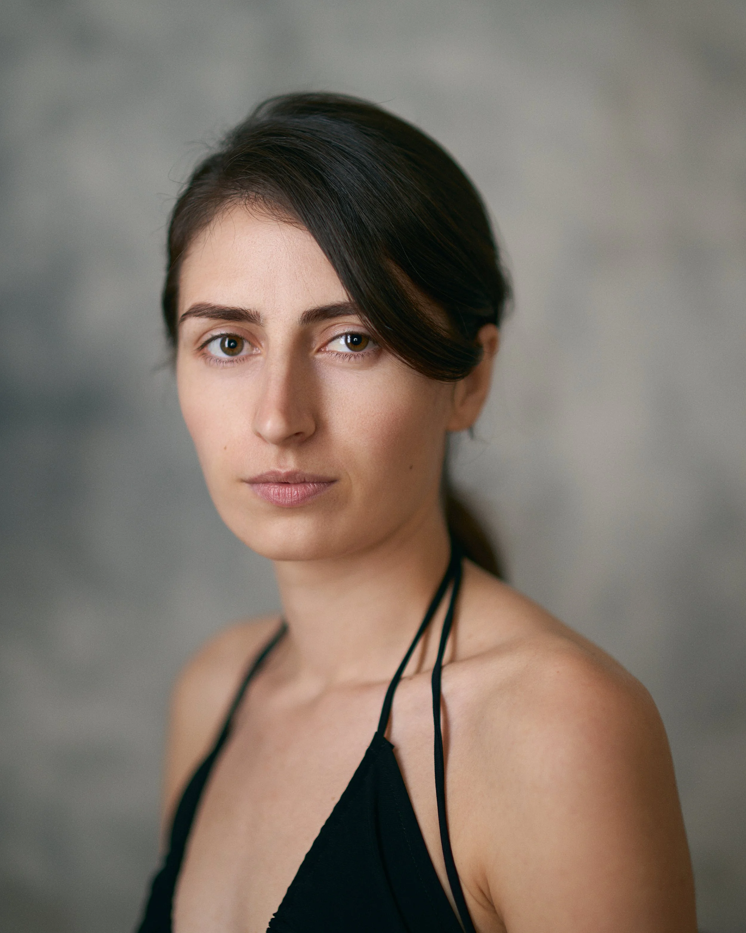 Close-up portrait of a woman with dark hair, light skin, wearing a black spaghetti strap top, against a blurred gray background.