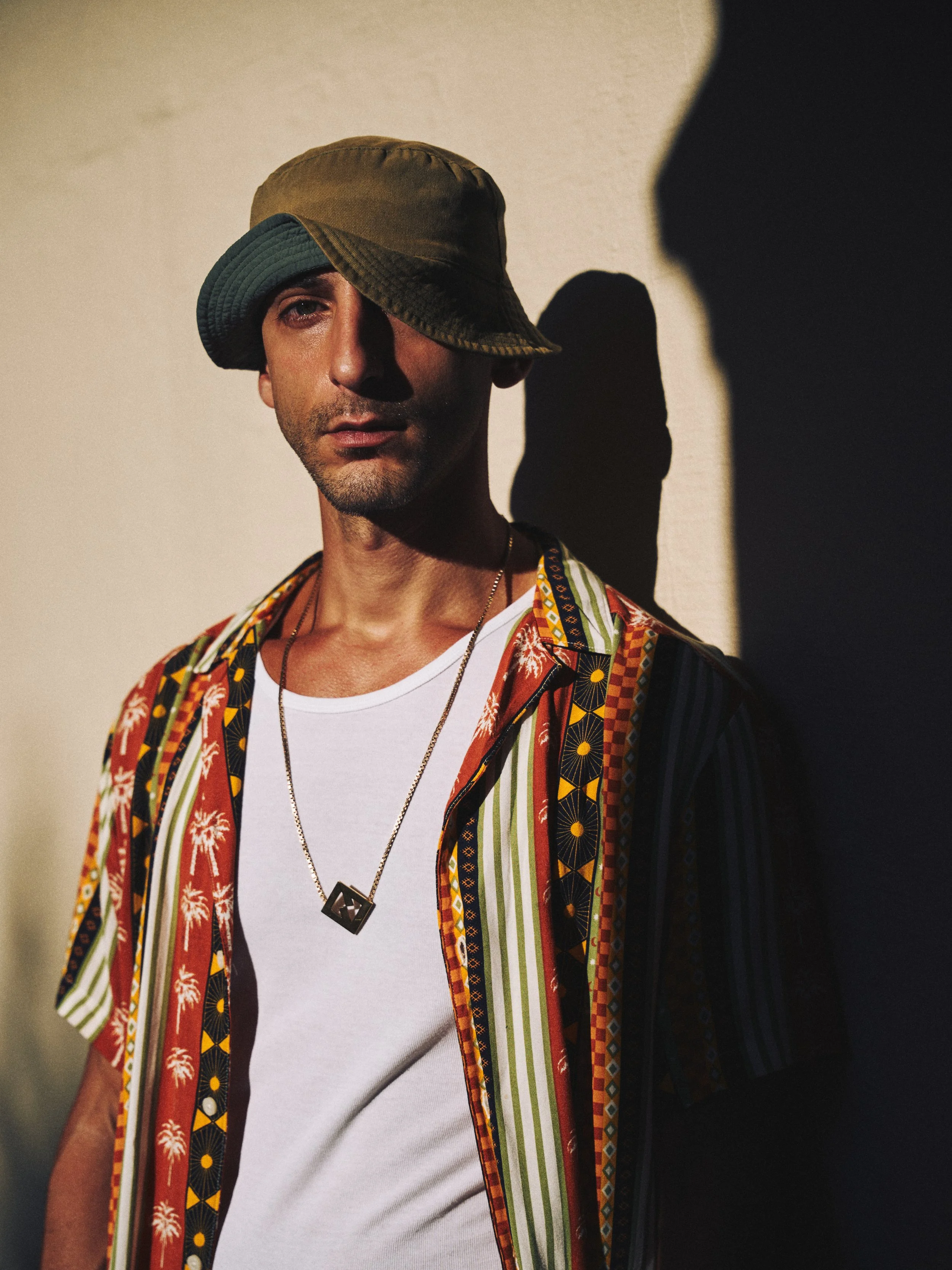 A man wearing a multicolored, patterned short-sleeve shirt, a white t-shirt, a gold necklace with a pendant, and a bucket hat layered with another hat, standing against a wall with a shadow cast behind him.