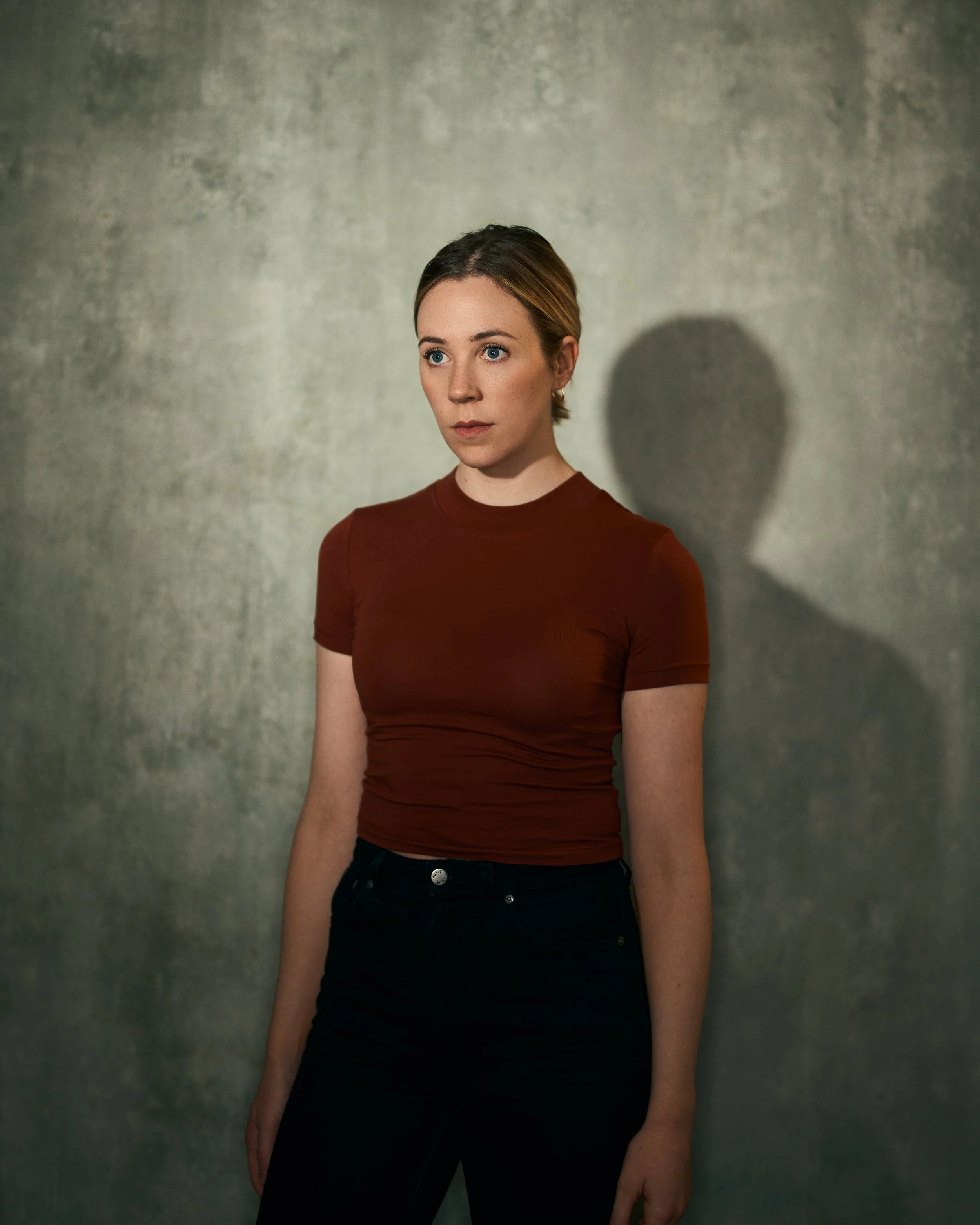 A young woman with short hair and blue eyes standing against a plain textured gray wall. She is wearing a fitted red t-shirt and dark pants, looking slightly to her left with a serious expression. Her shadow is cast to her right on the wall.