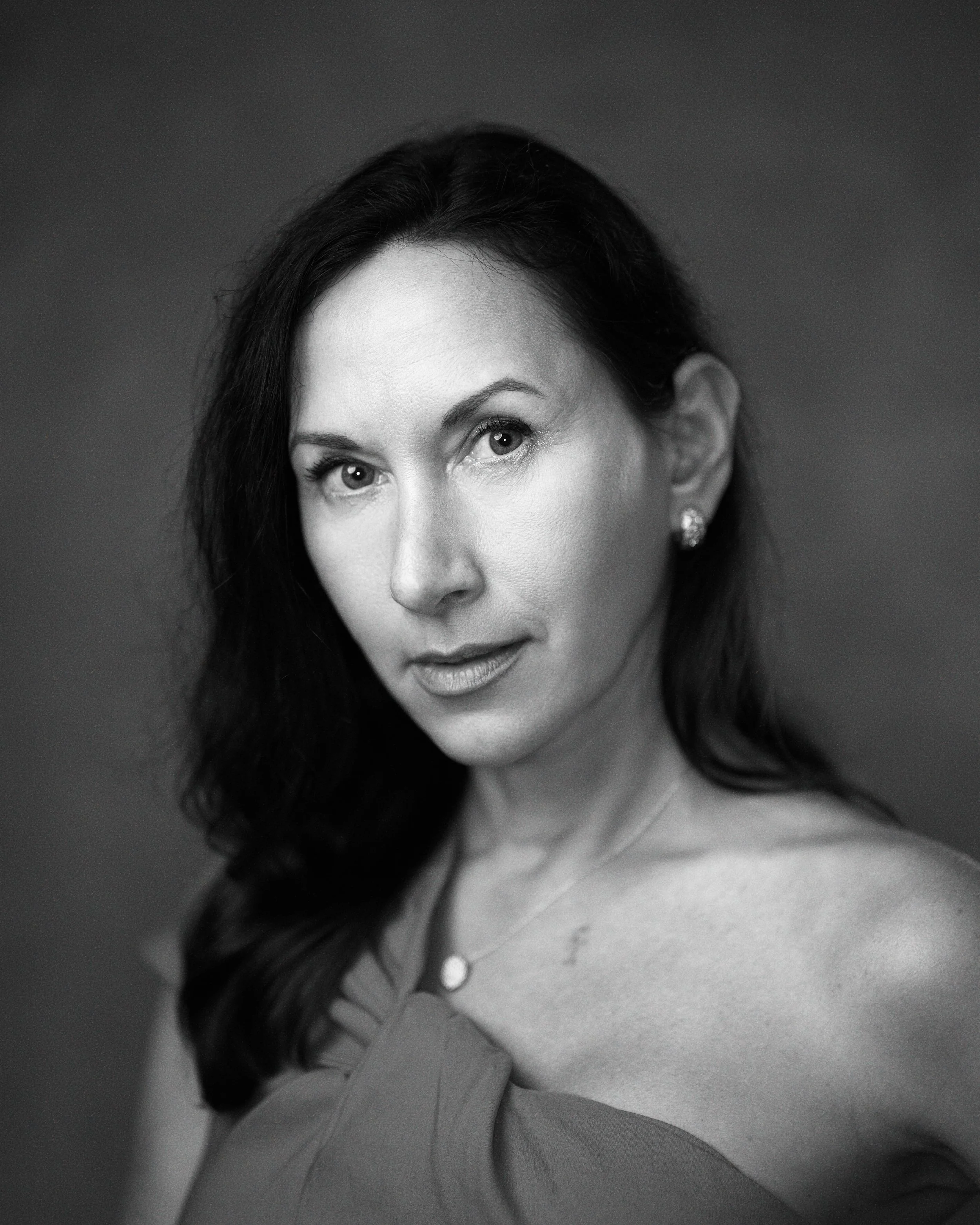 A black and white close-up portrait of a woman with dark hair, wearing earrings and a necklace, looking at the camera with a slight smile.