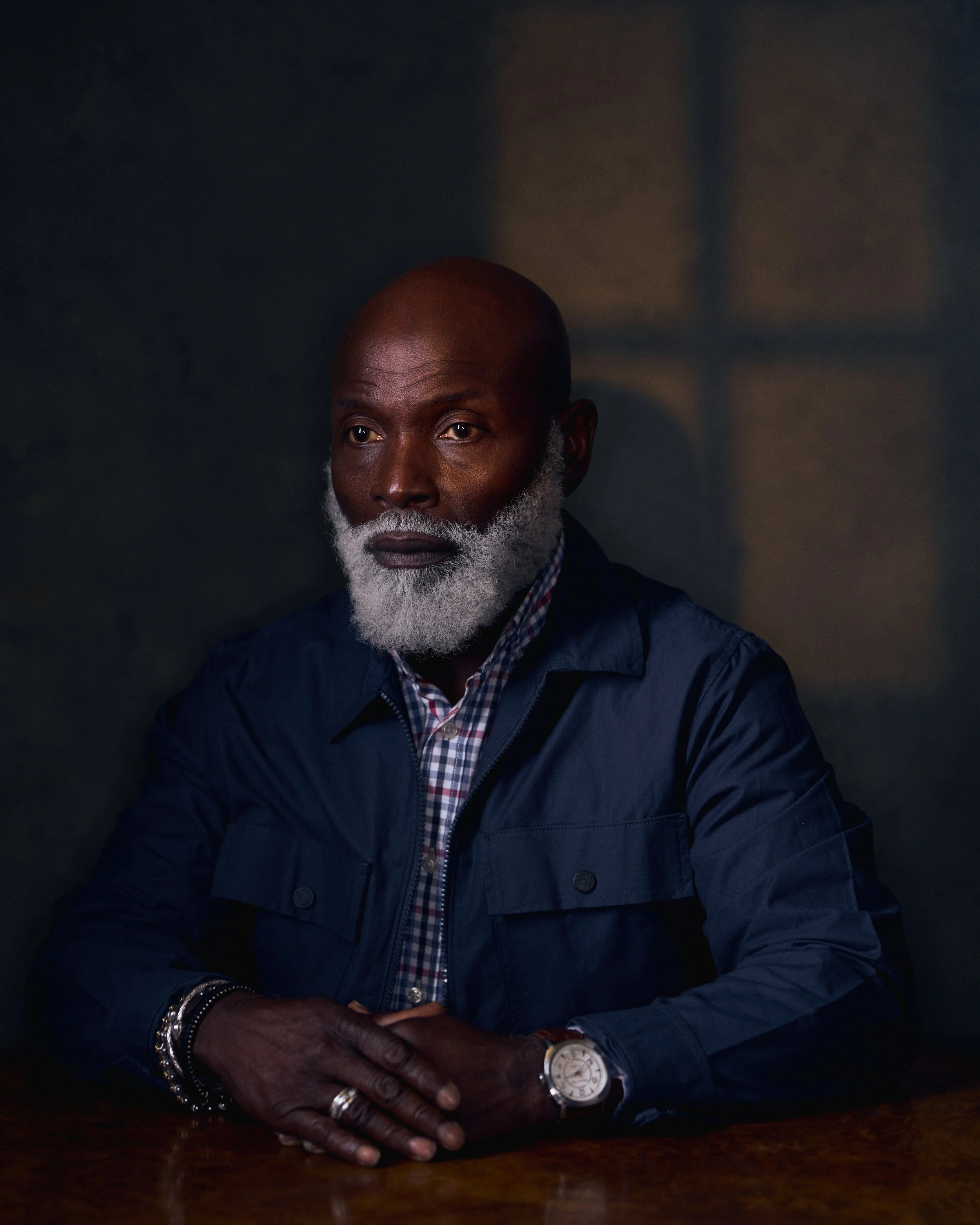 A portrait of an older African American man with a bald head and gray beard, wearing a navy blue jacket over a checkered shirt, sitting at a dark wooden table with his hands clasped, in a dimly lit room.