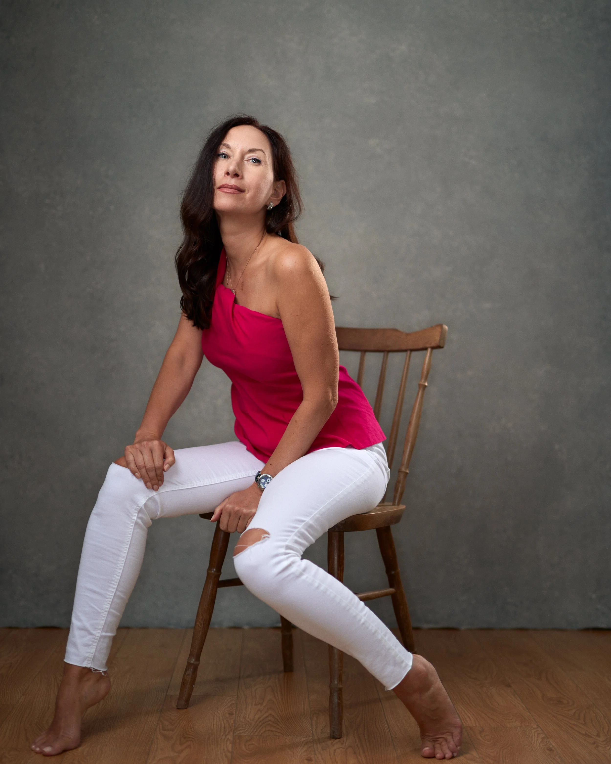 A woman with long dark hair, wearing a pink off-the-shoulder top and white ripped jeans, sitting barefoot on a wooden chair against a plain gray background.