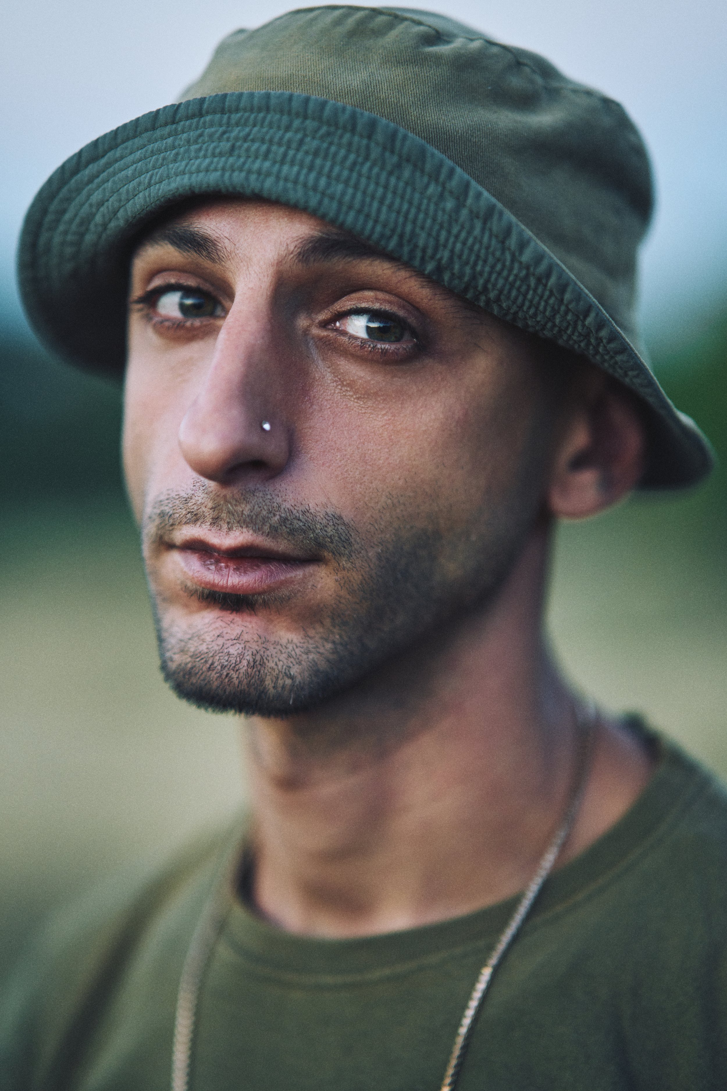 Close-up photo of a man wearing a green bucket hat and a green shirt, with a nose piercing, looking at the camera with a neutral expression.