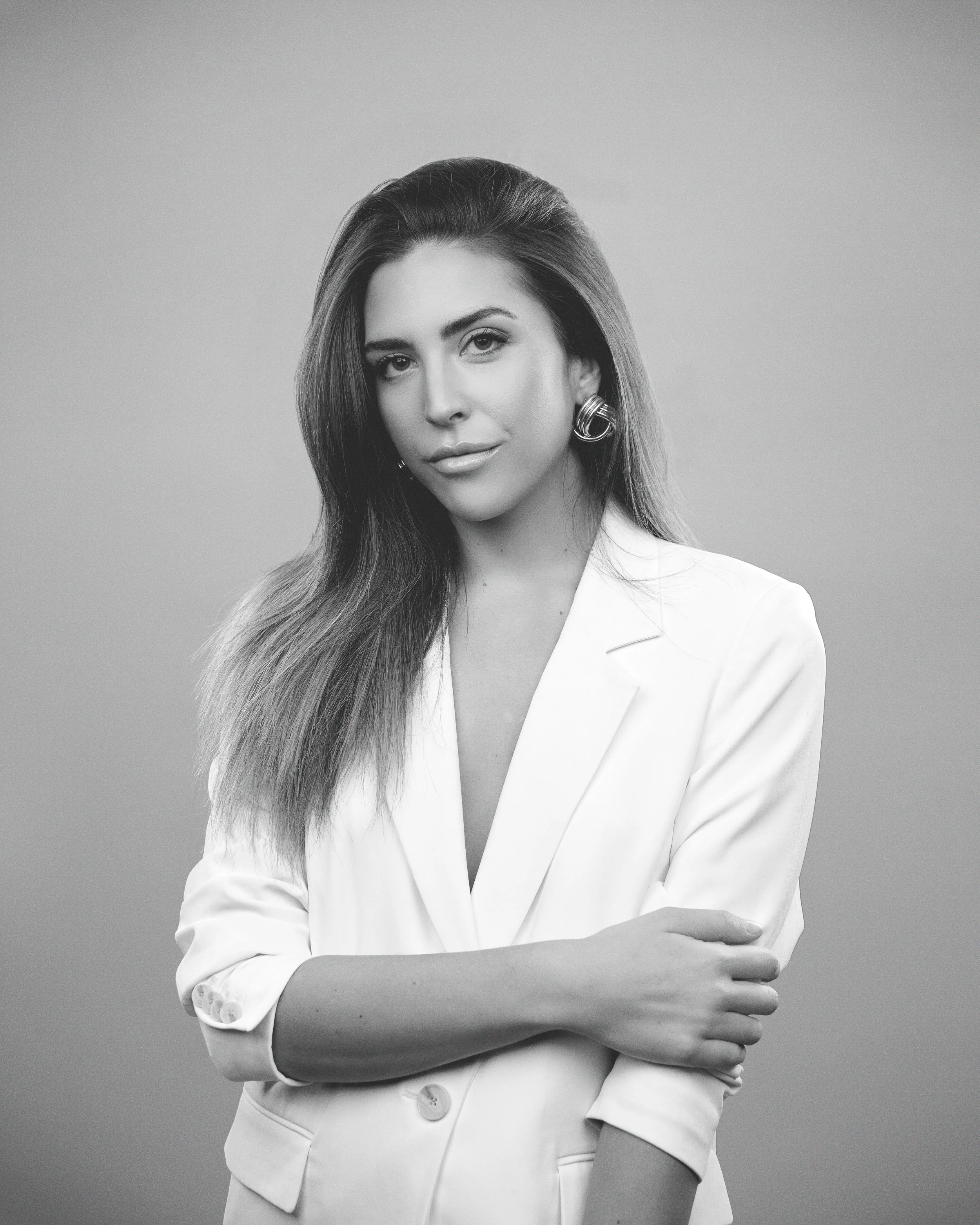 Black and white portrait of a woman with long hair, wearing a white blazer and large hoop earrings, looking directly at the camera with a confident expression.