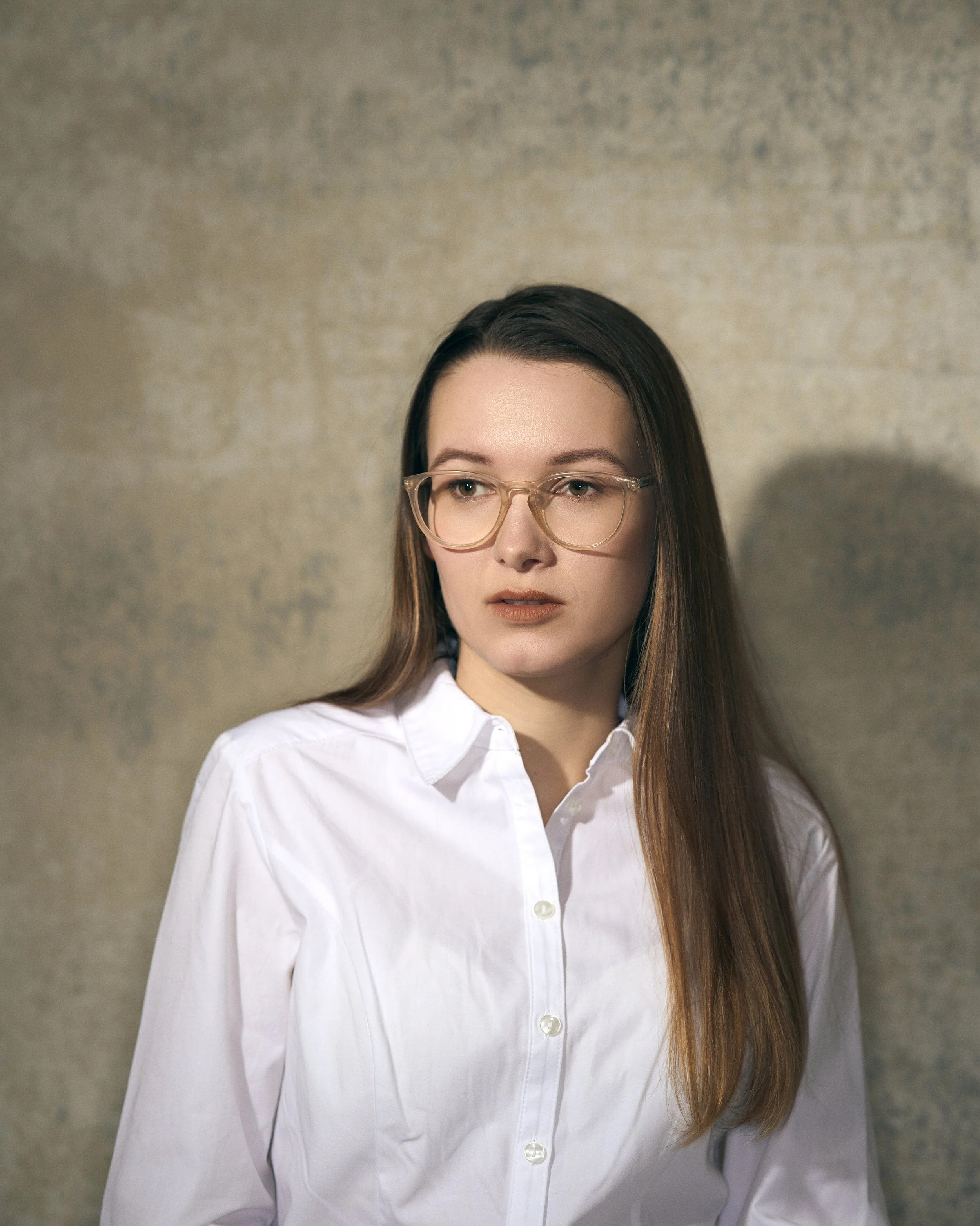 A young woman with long brown hair, wearing clear glasses and a white button-up shirt, standing against a textured beige wall.