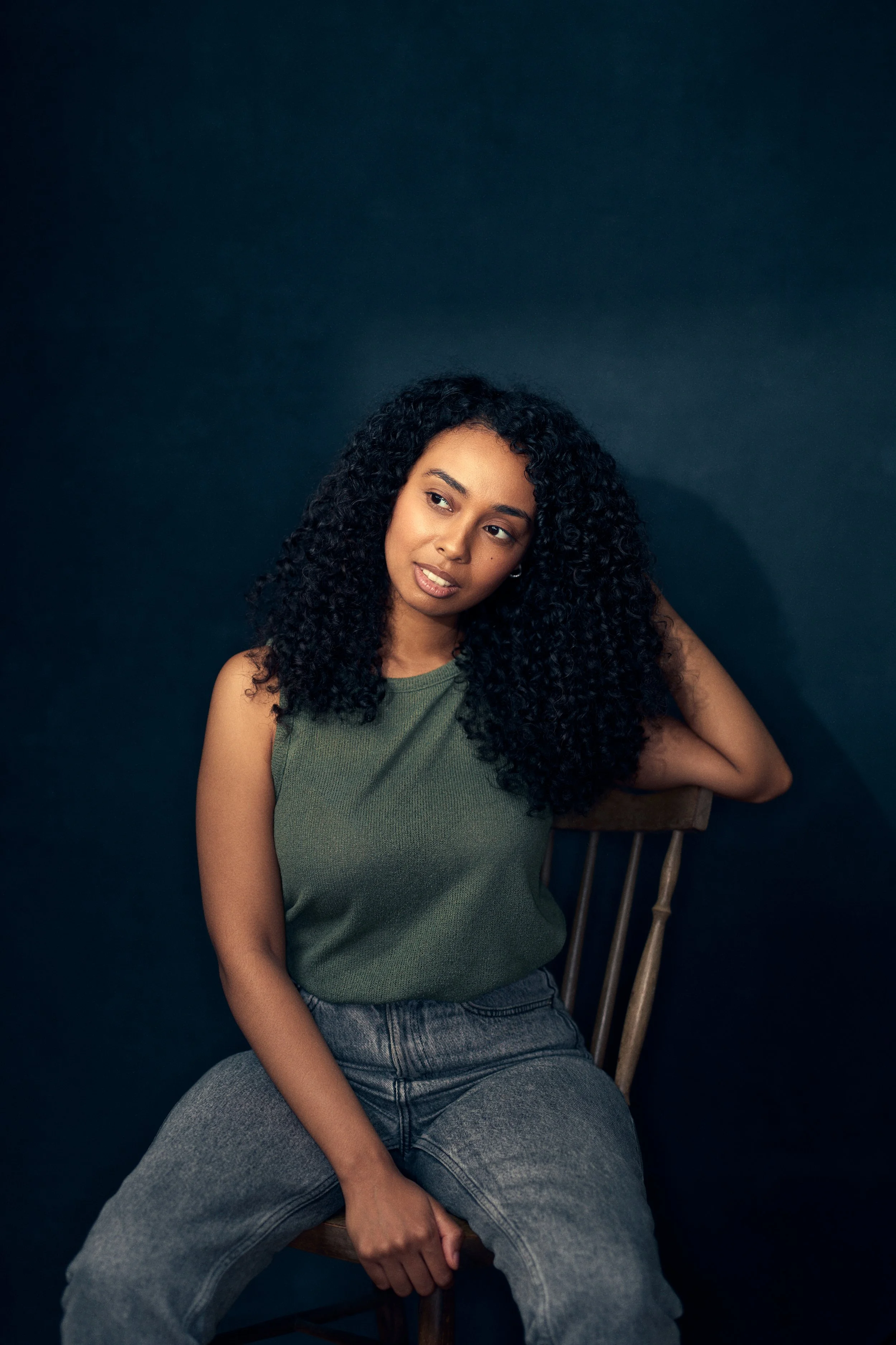 A young woman with curly black hair, wearing a green sleeveless top and gray jeans, sitting on a wooden chair against a dark background.