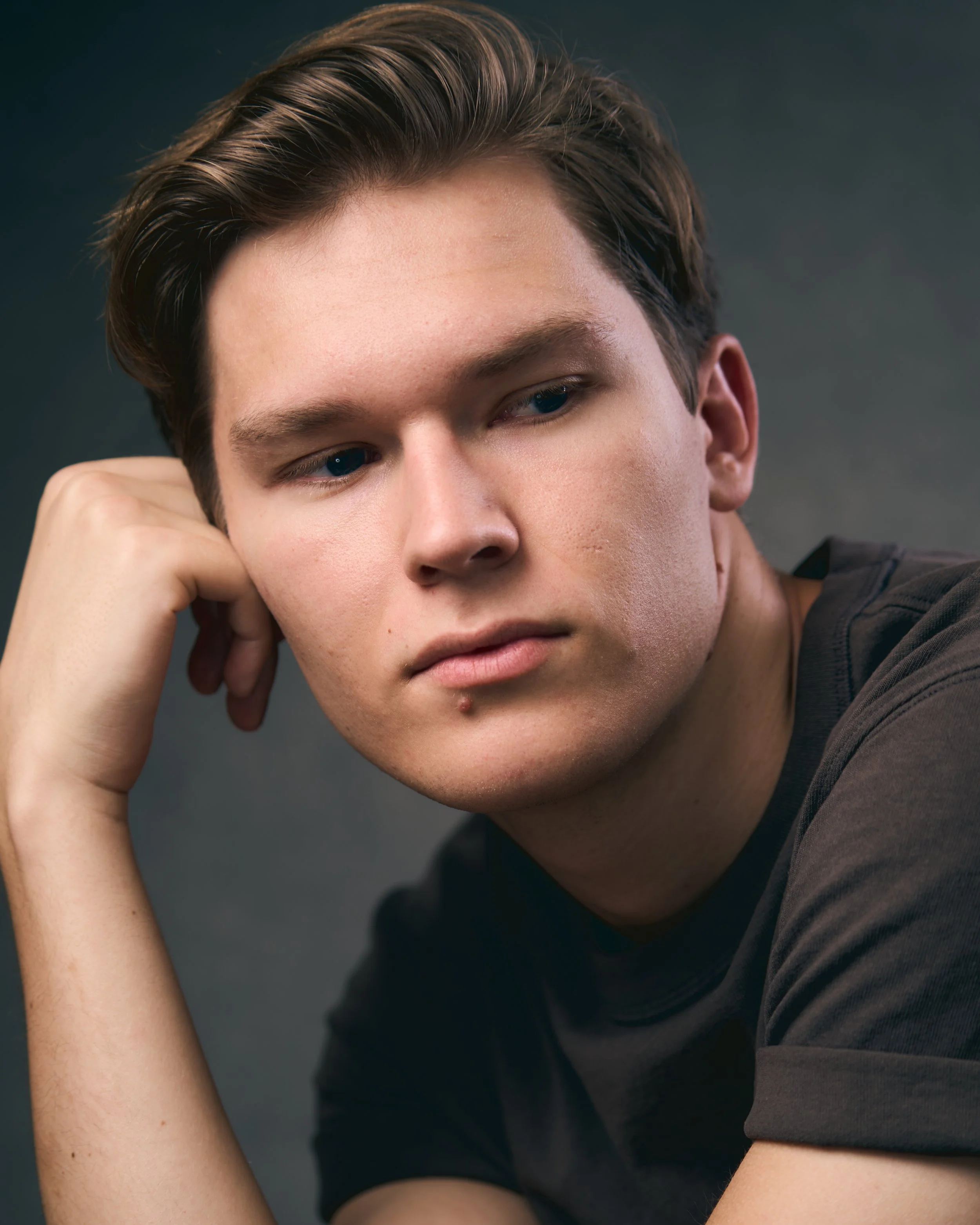 A young man with brown hair resting his head on his left hand, looking thoughtful, wearing a black shirt against a dark background.