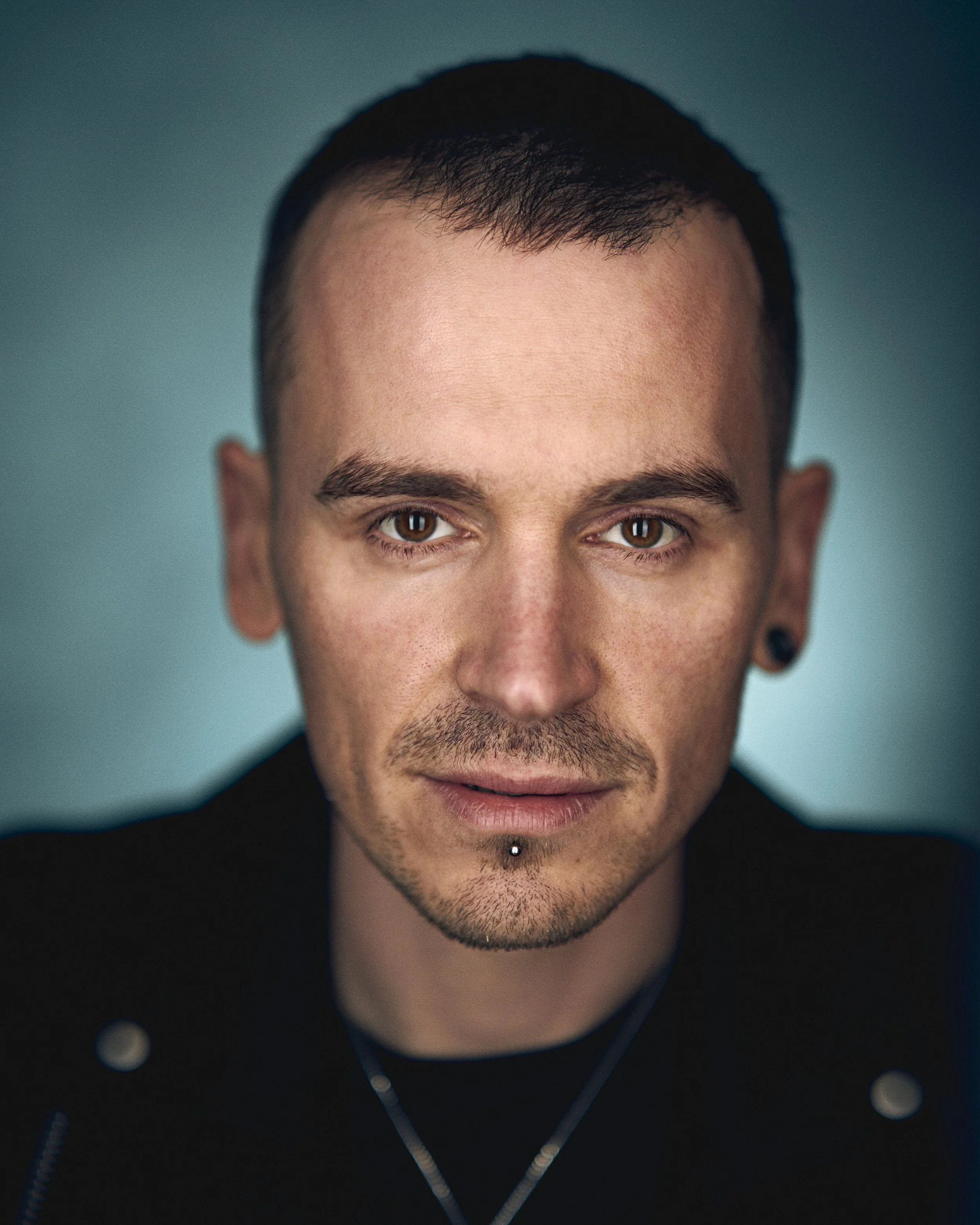 Close-up portrait of a young man with brown eyes, short dark hair, light facial hair, and ear gauges, wearing a black jacket and a silver necklace, against a blurred teal background.